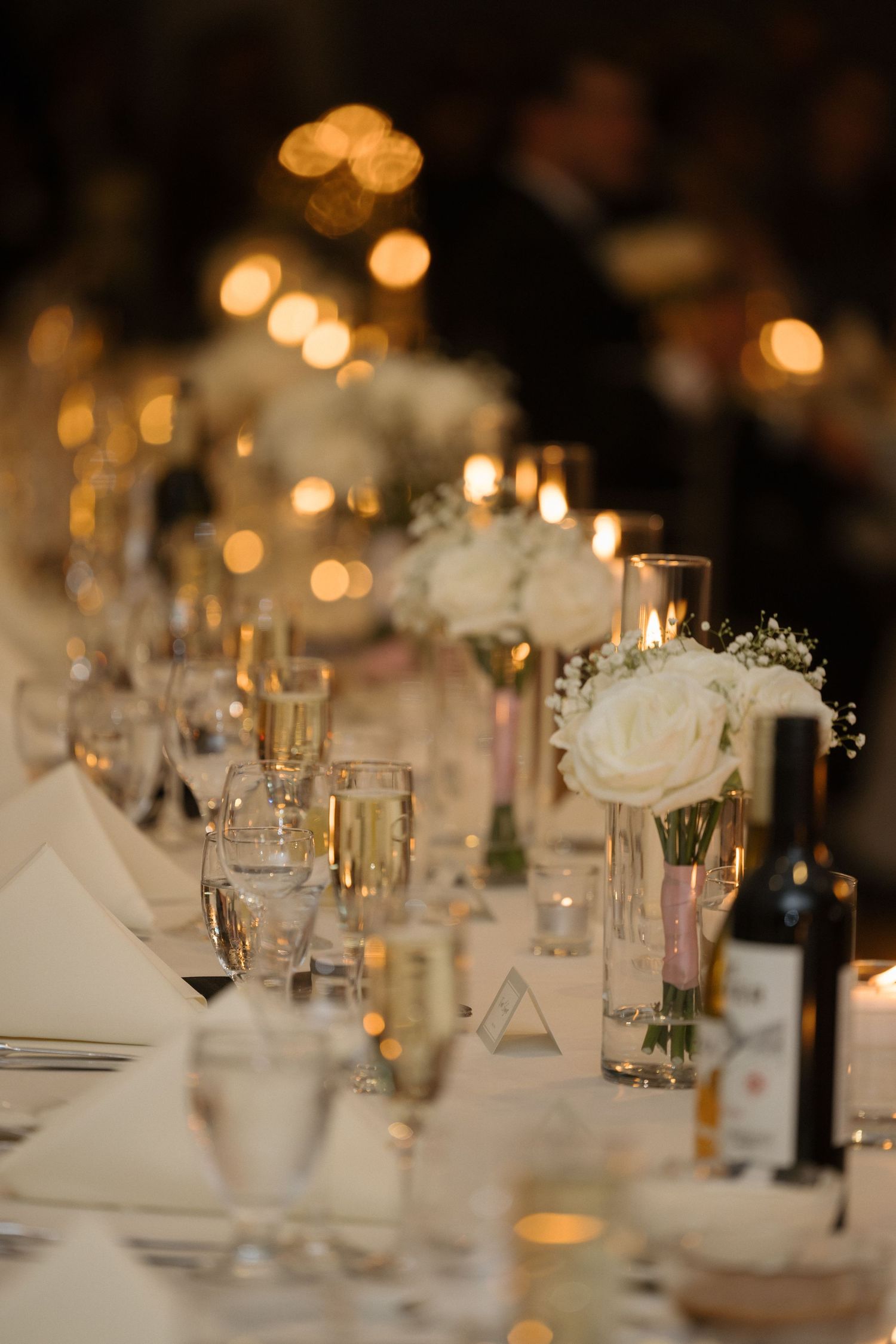 Elegant wedding reception table with white rose centerpieces, candles, and champagne glasses.