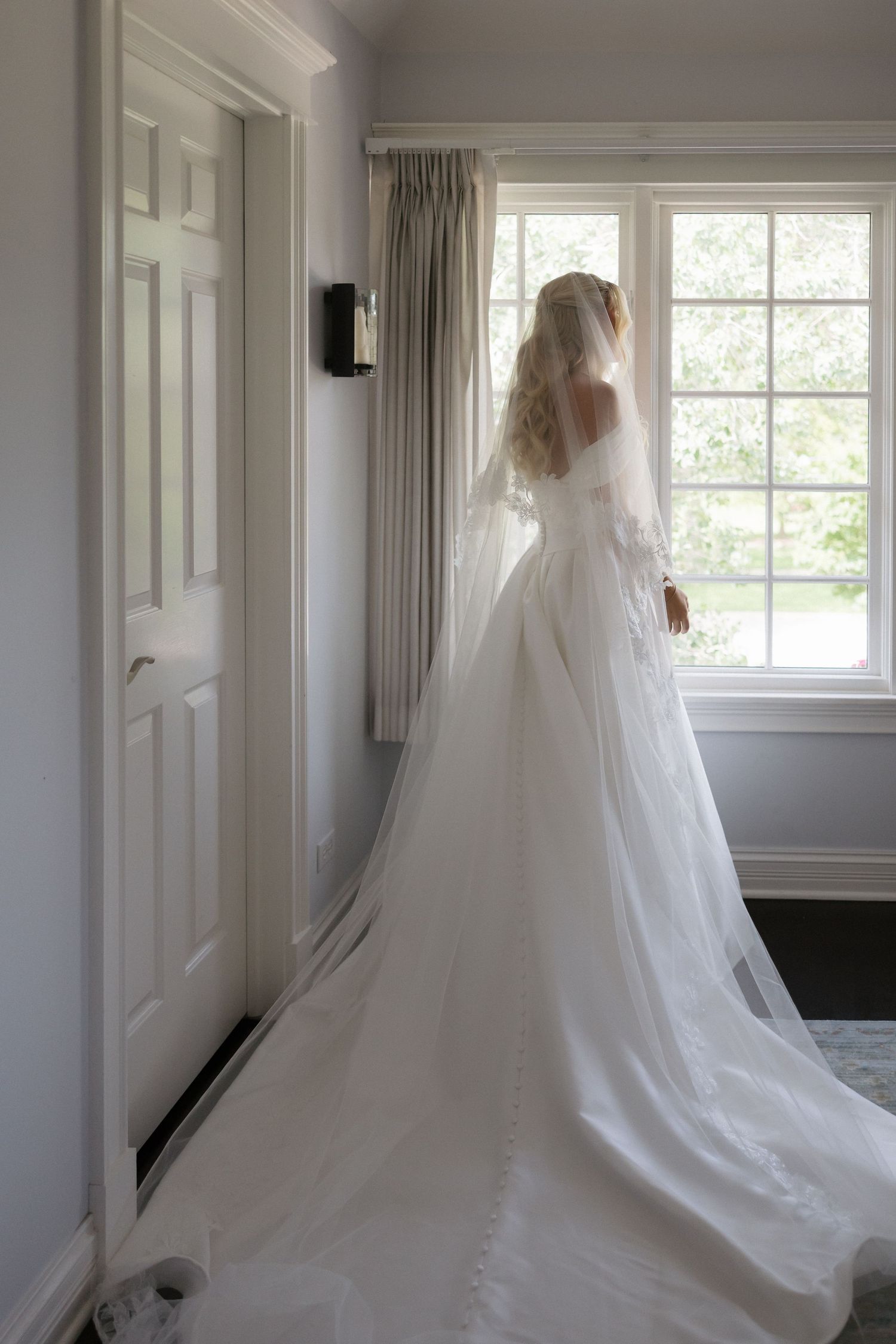 Bride in elegant white wedding gown with long veil and train stands by window in soft natural light.
