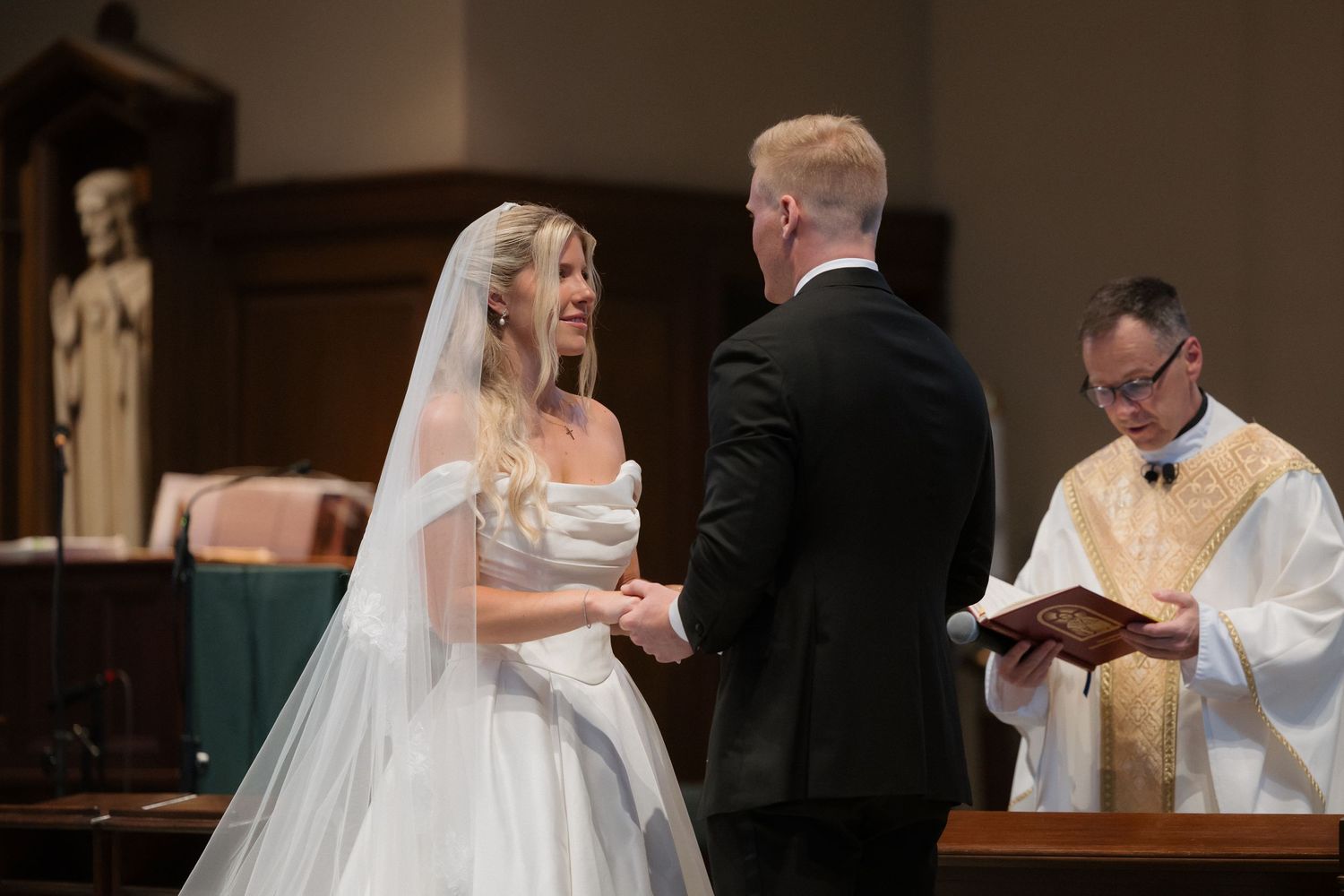 Bride and groom exchange vows at church altar, officiant holding book, bride in white gown and veil smiling.