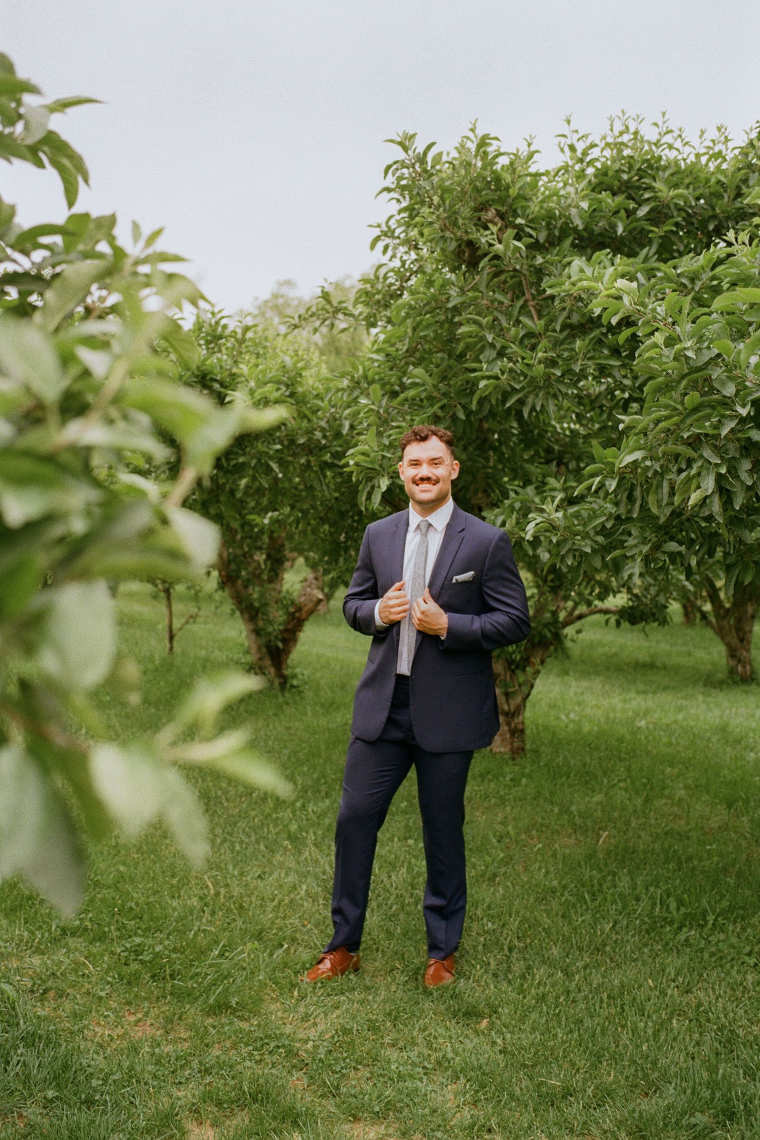 Smiling groom in navy suit with light blue tie standing in lush green orchard on wedding day.