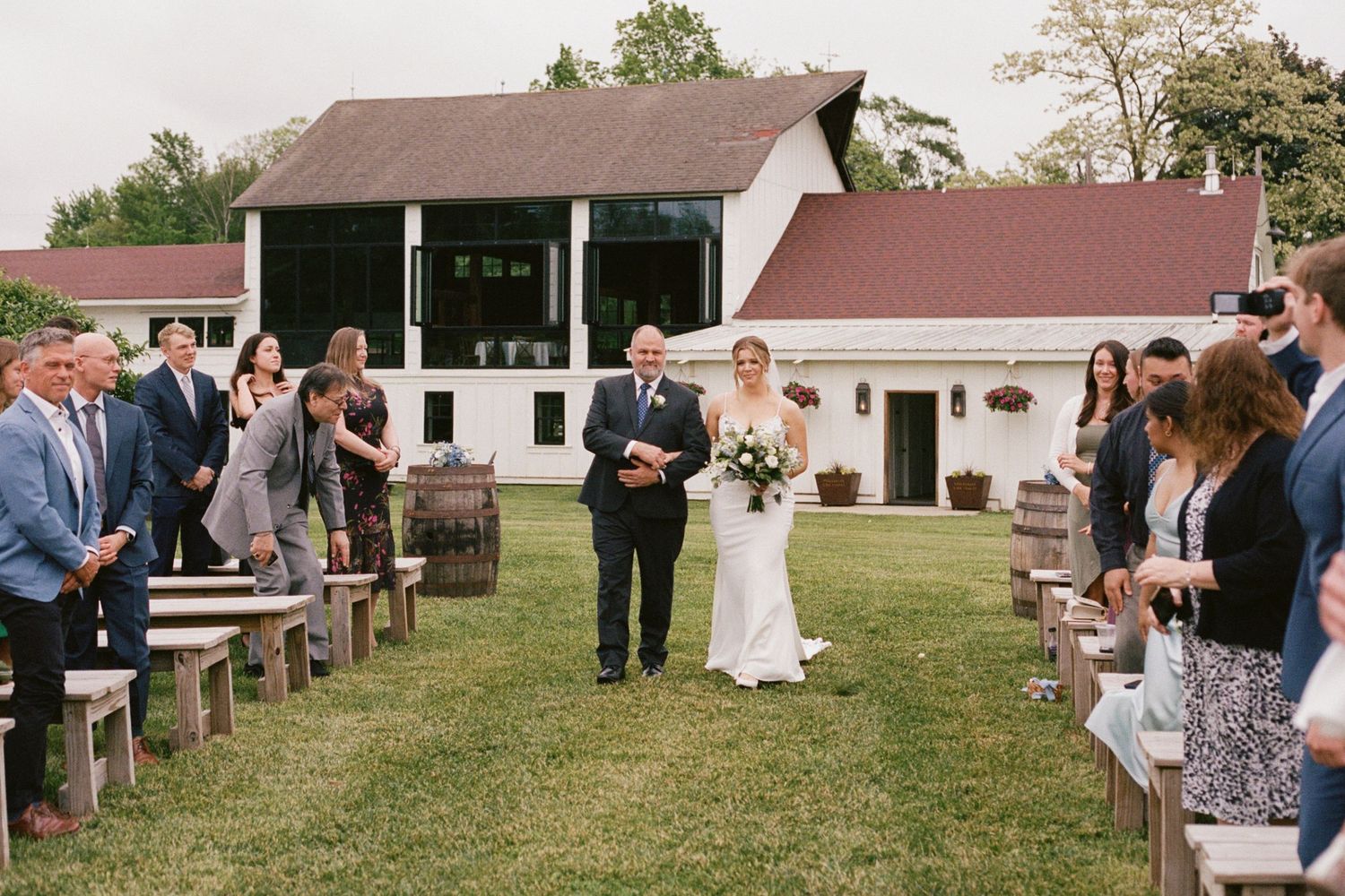 Bride walking down outdoor aisle with escort past wooden benches toward white barn wedding venue.