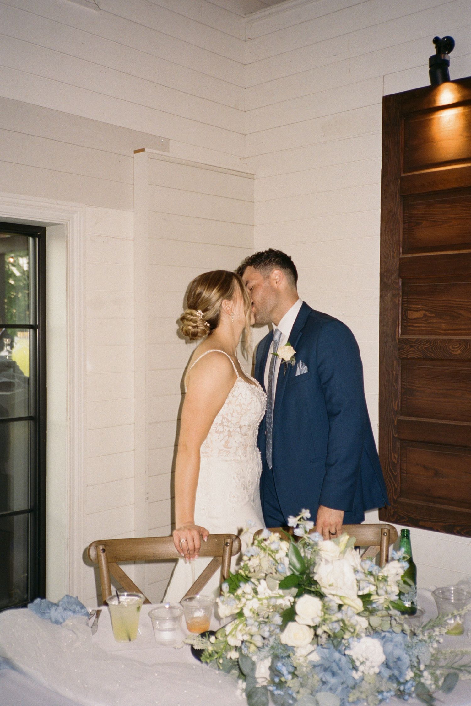 Bride and groom sharing a kiss at their wedding reception sweetheart table adorned with blue and white floral arrangements.