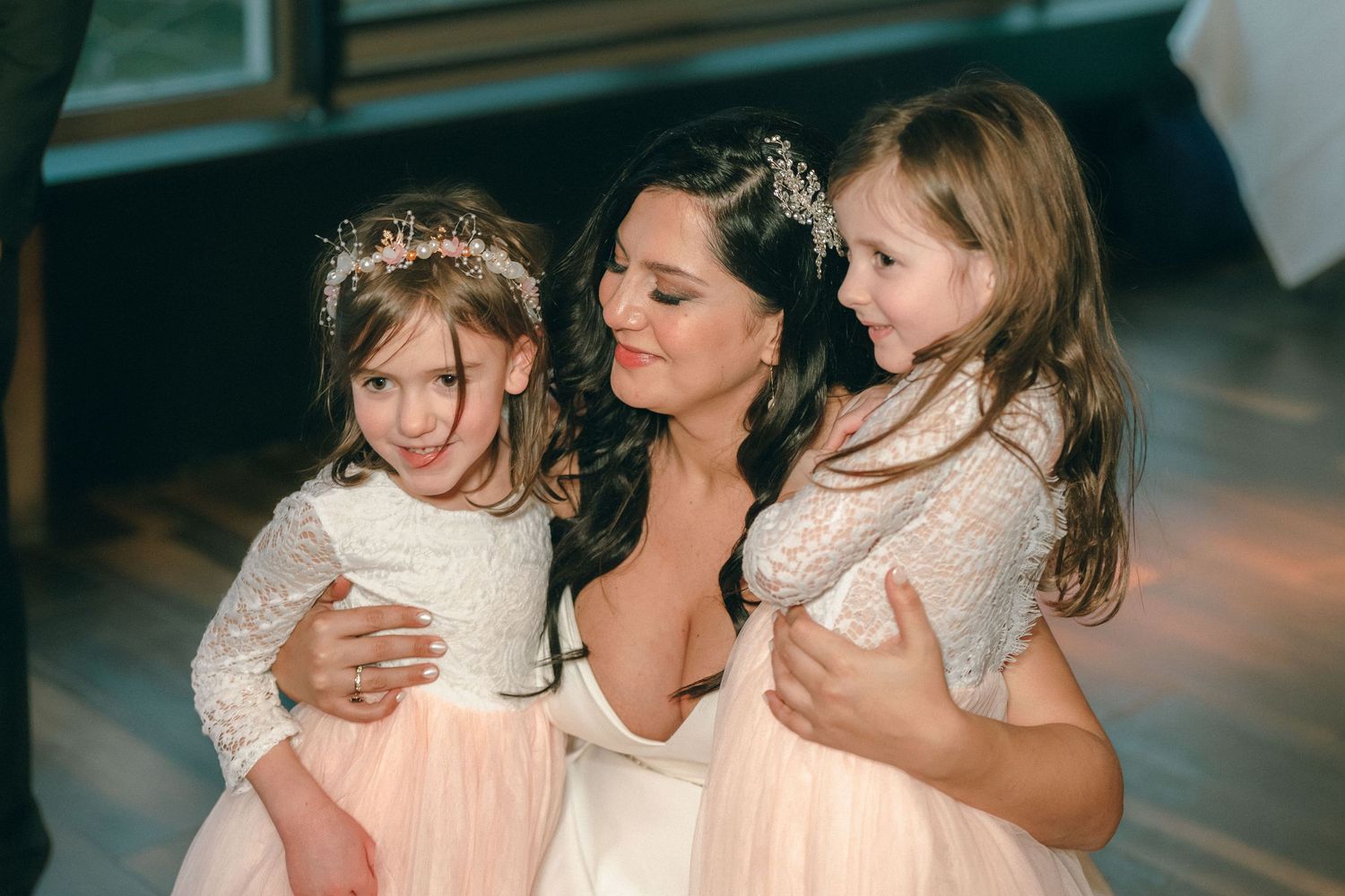 Beautiful bride hugs two young flower girls in pink tulle dresses at wedding.
