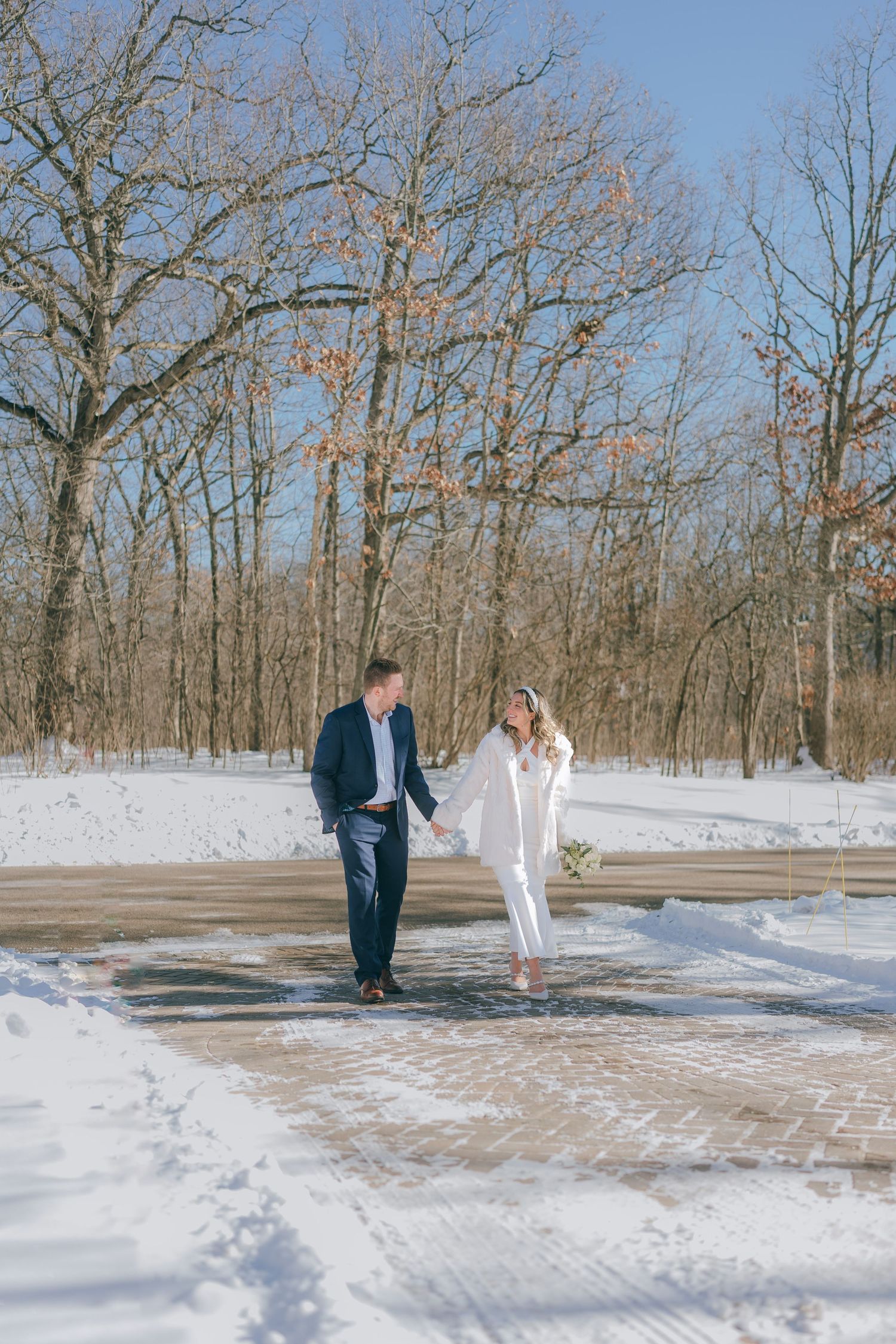Couple holding hands walking on snowy brick path surrounded by bare winter trees on sunny day.