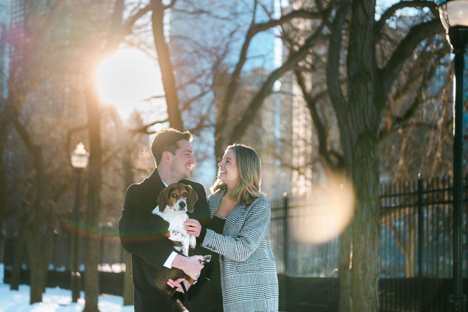 Couple holding their small dog together in a sunlit snowy park with bare trees.