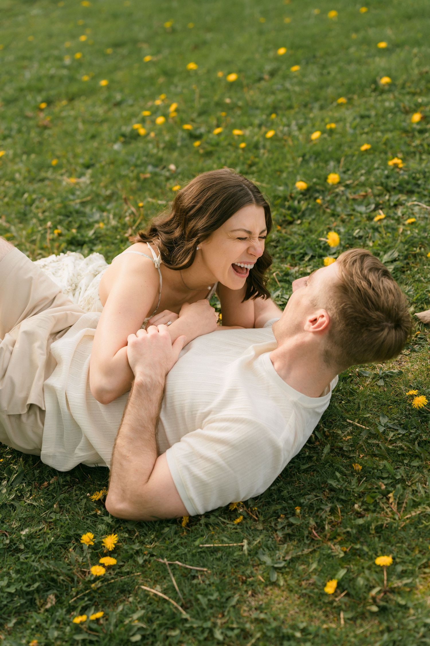 A happy couple laughs and plays together while lying on a grass field with dandelions.