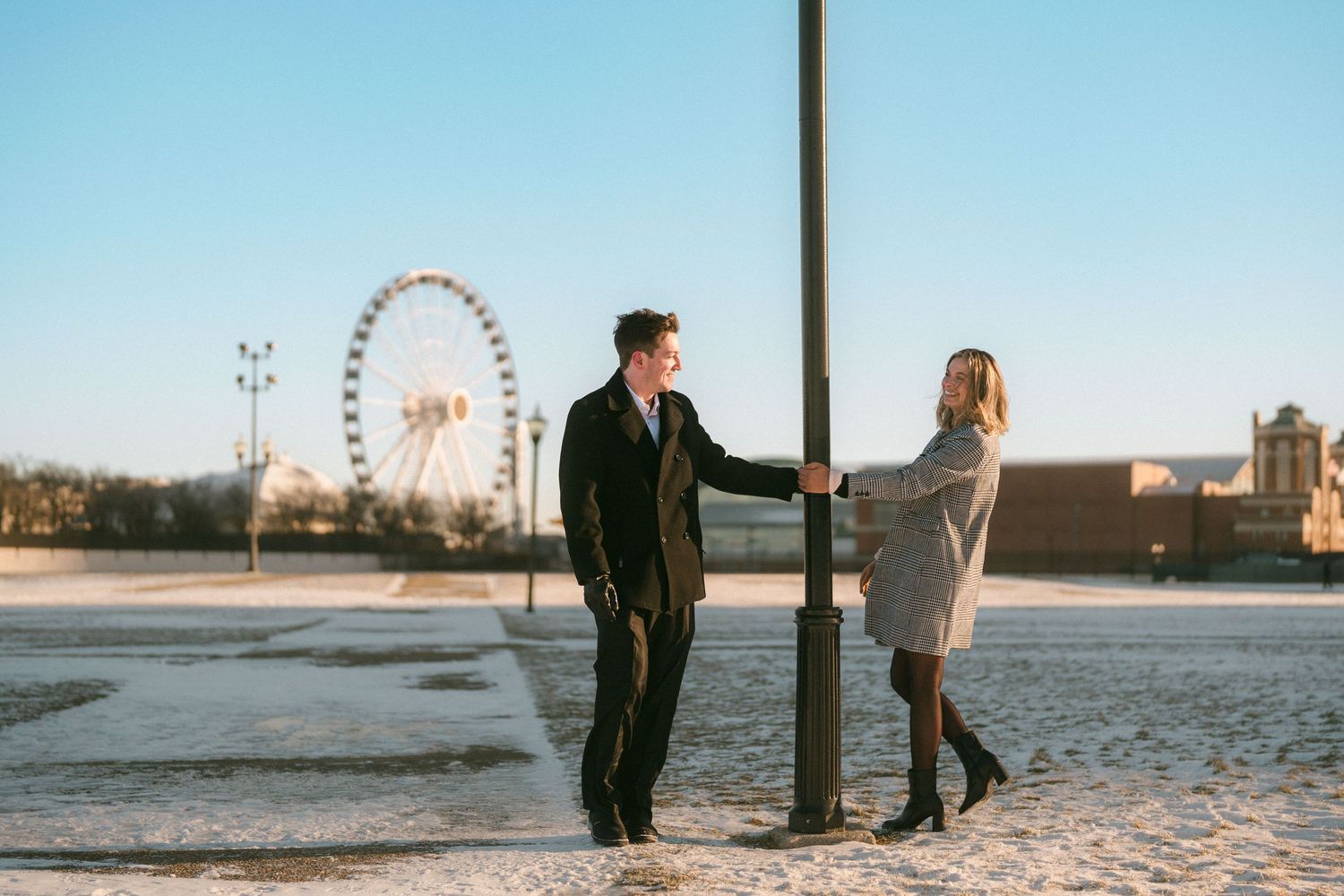 Couple playfully posing around a lamppost with a ferris wheel and snowy ground in the background.