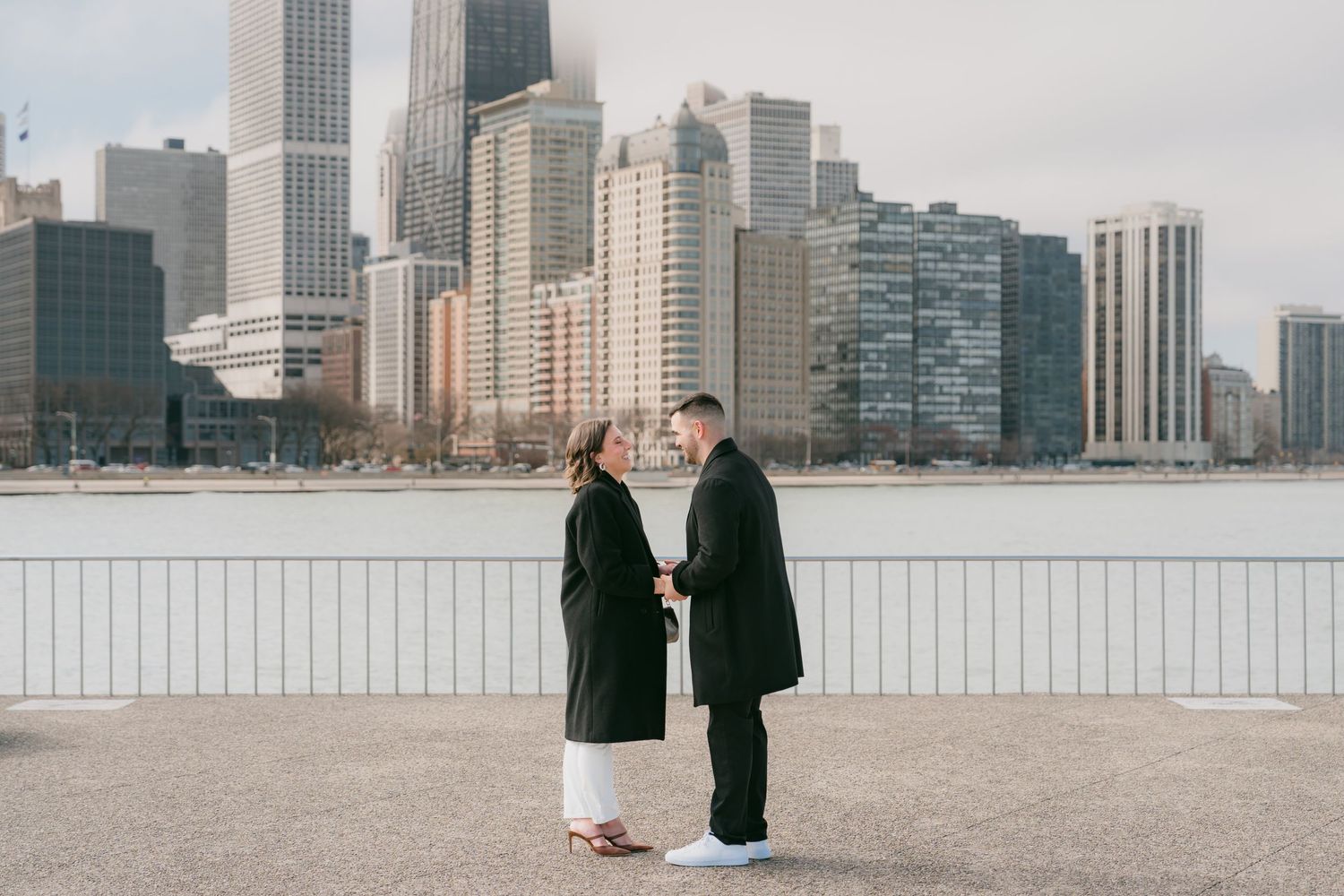Romantic couple holding hands facing each other on Chicago lakefront with winter city skyline backdrop.