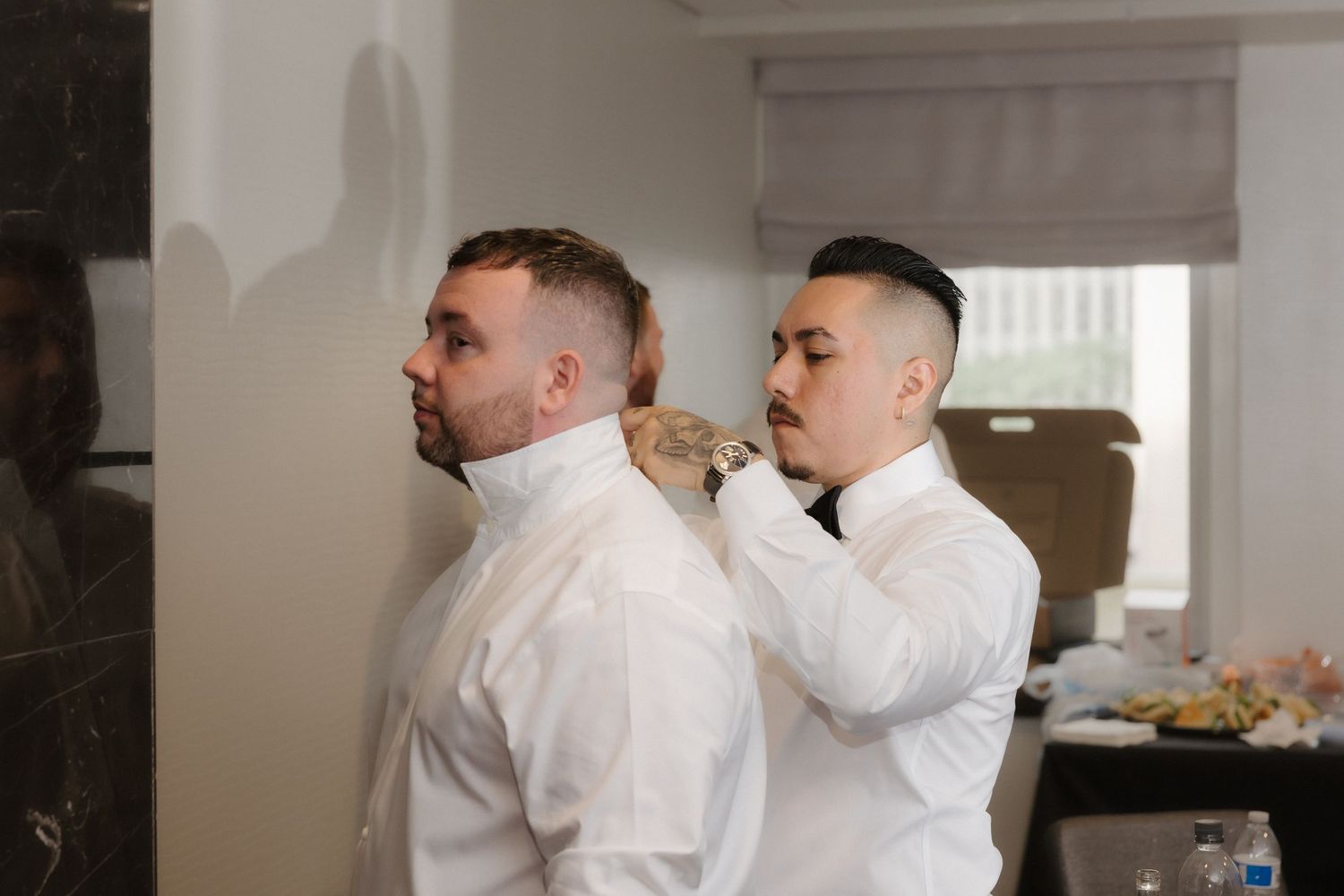A man in a white shirt helps another adjust his collar and tie during wedding preparations.