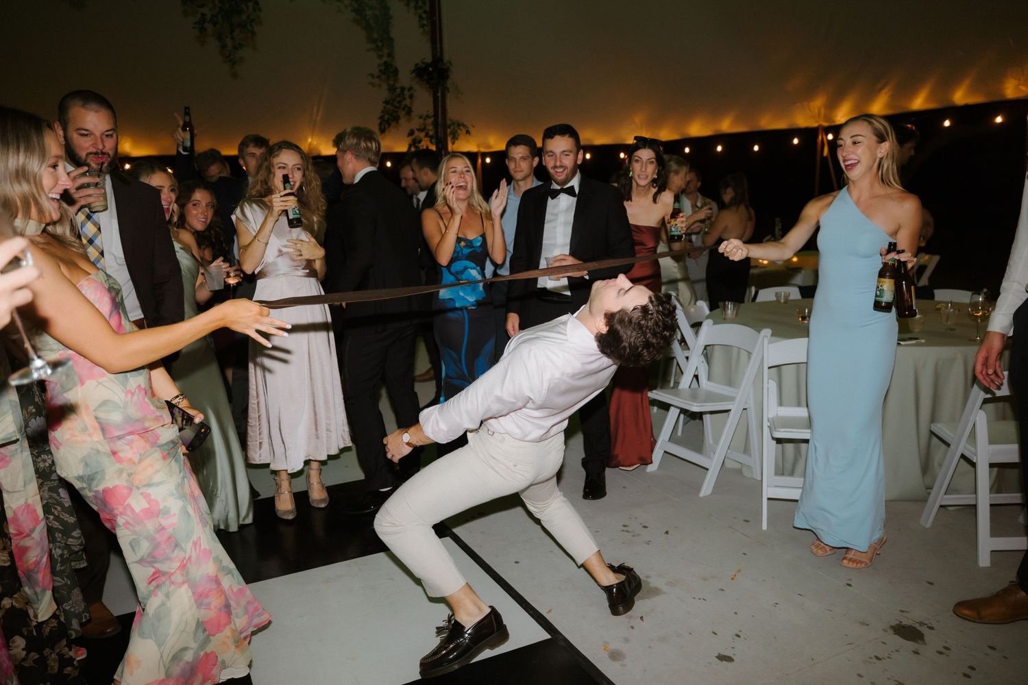 Wedding guest performs energetic dance moves on checkered floor as guests cheer at outdoor tented reception.