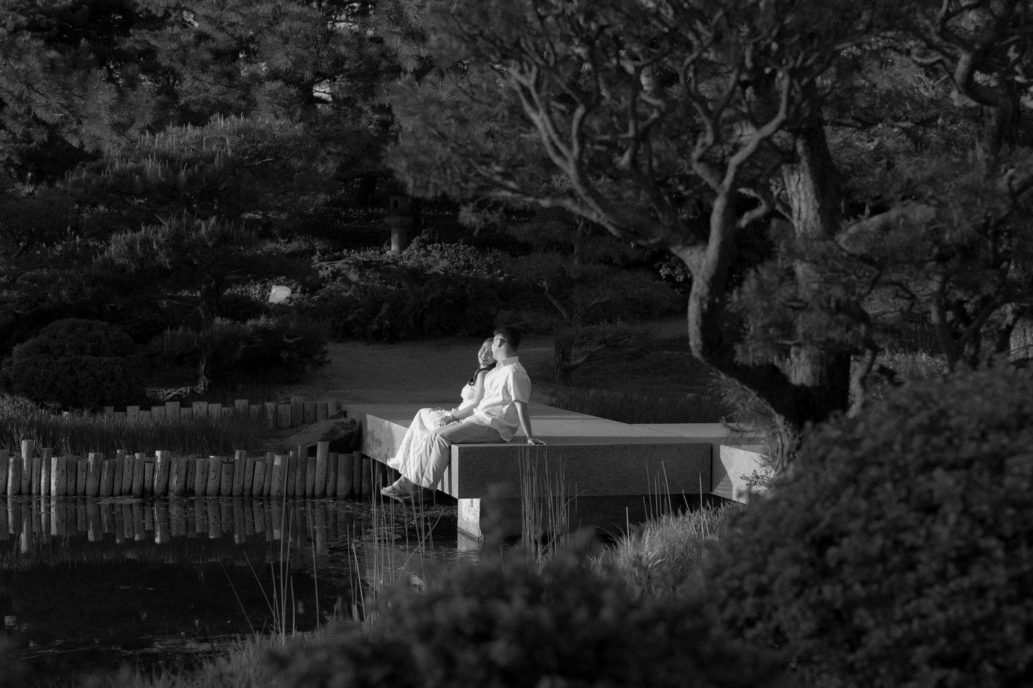 Black and white photo of a person in white dress sitting on a stone bench in a serene Japanese garden.