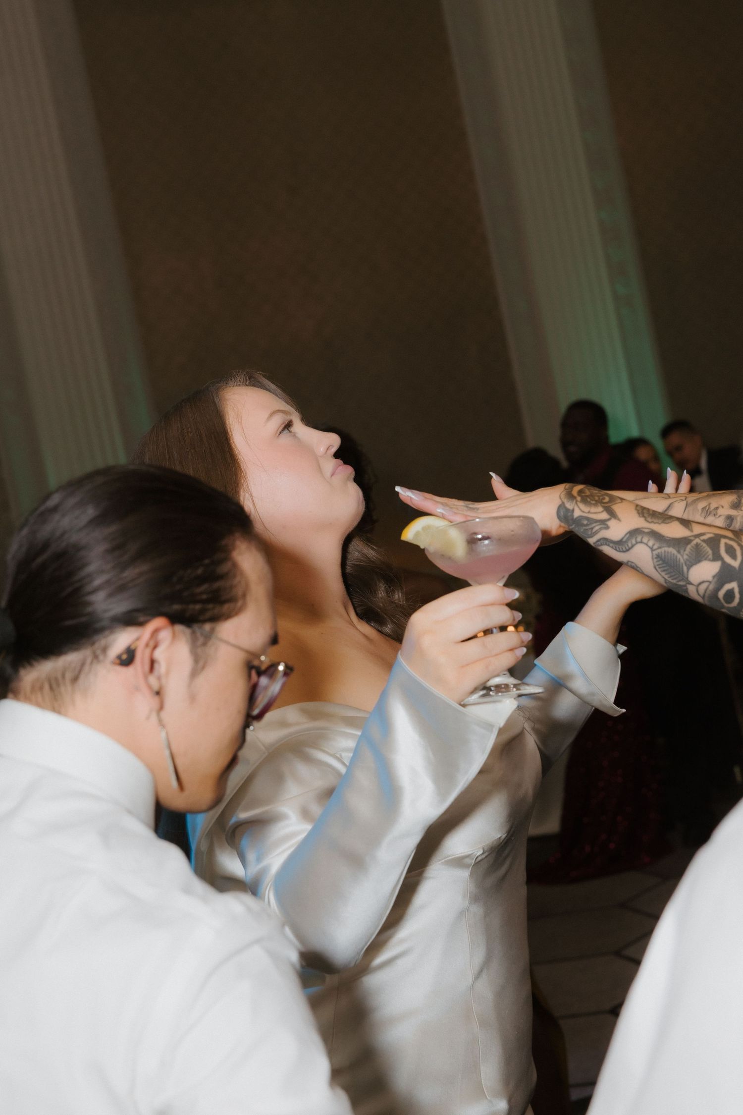 Woman in white satin dress holding pink cocktail at wedding reception, reaching toward tattooed hand.