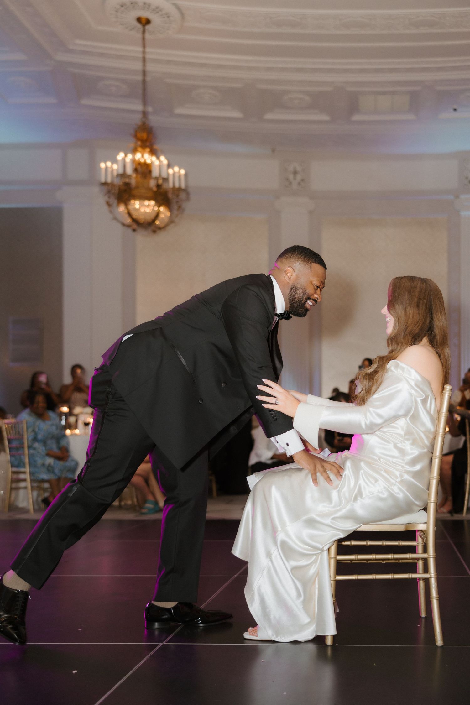 Groom in black tuxedo bows toward bride in white satin gown seated on chair during wedding reception dance.