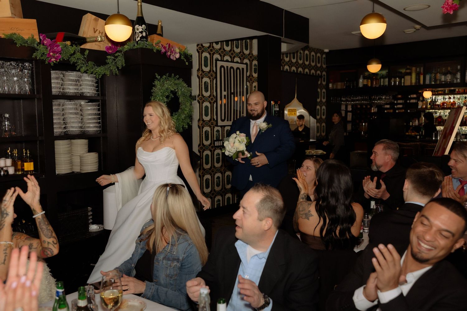 Bride and groom make grand entrance at wedding reception in elegant restaurant venue with cheering guests.