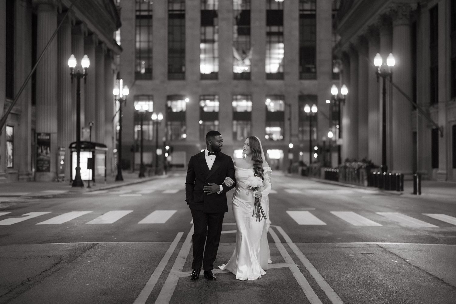Black and white photo of a bride and groom standing in a crosswalk at night in an illuminated city street.