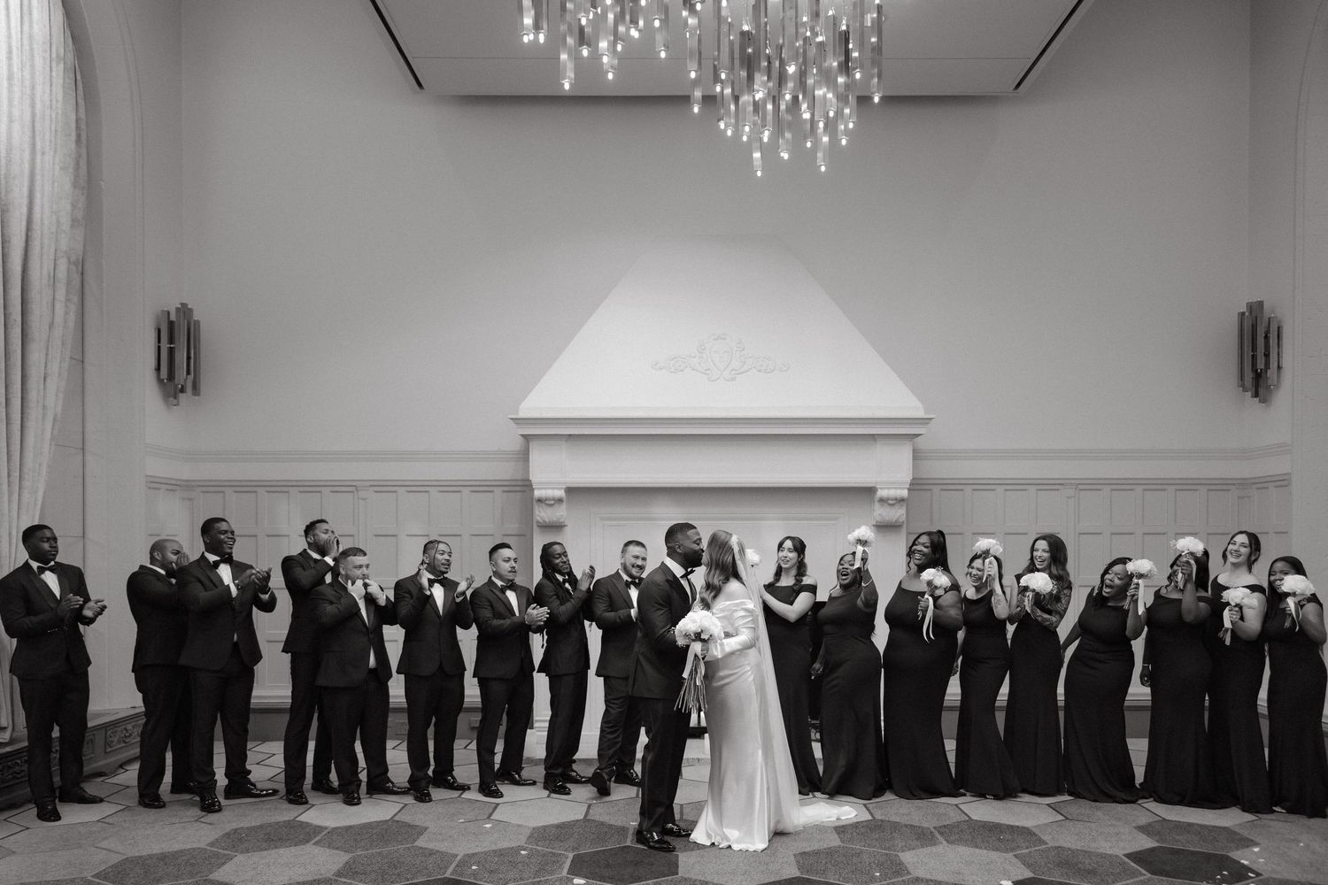 Black and white wedding photo of couple sharing first dance surrounded by bridal party in elegant ballroom with chandelier.