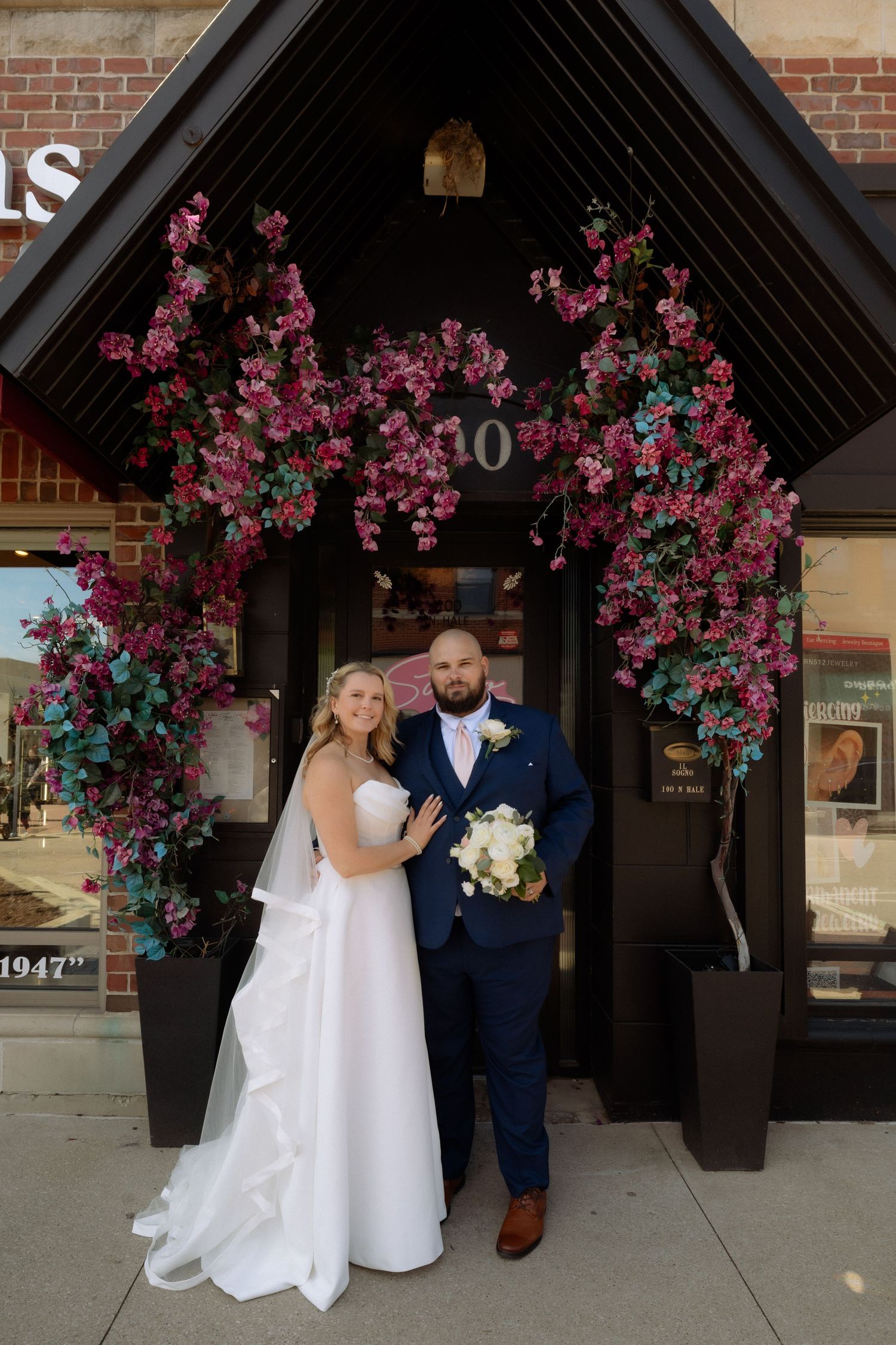 Bride and groom pose under floral arch with pink blooms at building entrance numbered 10, groom in navy suit, bride in white gown.