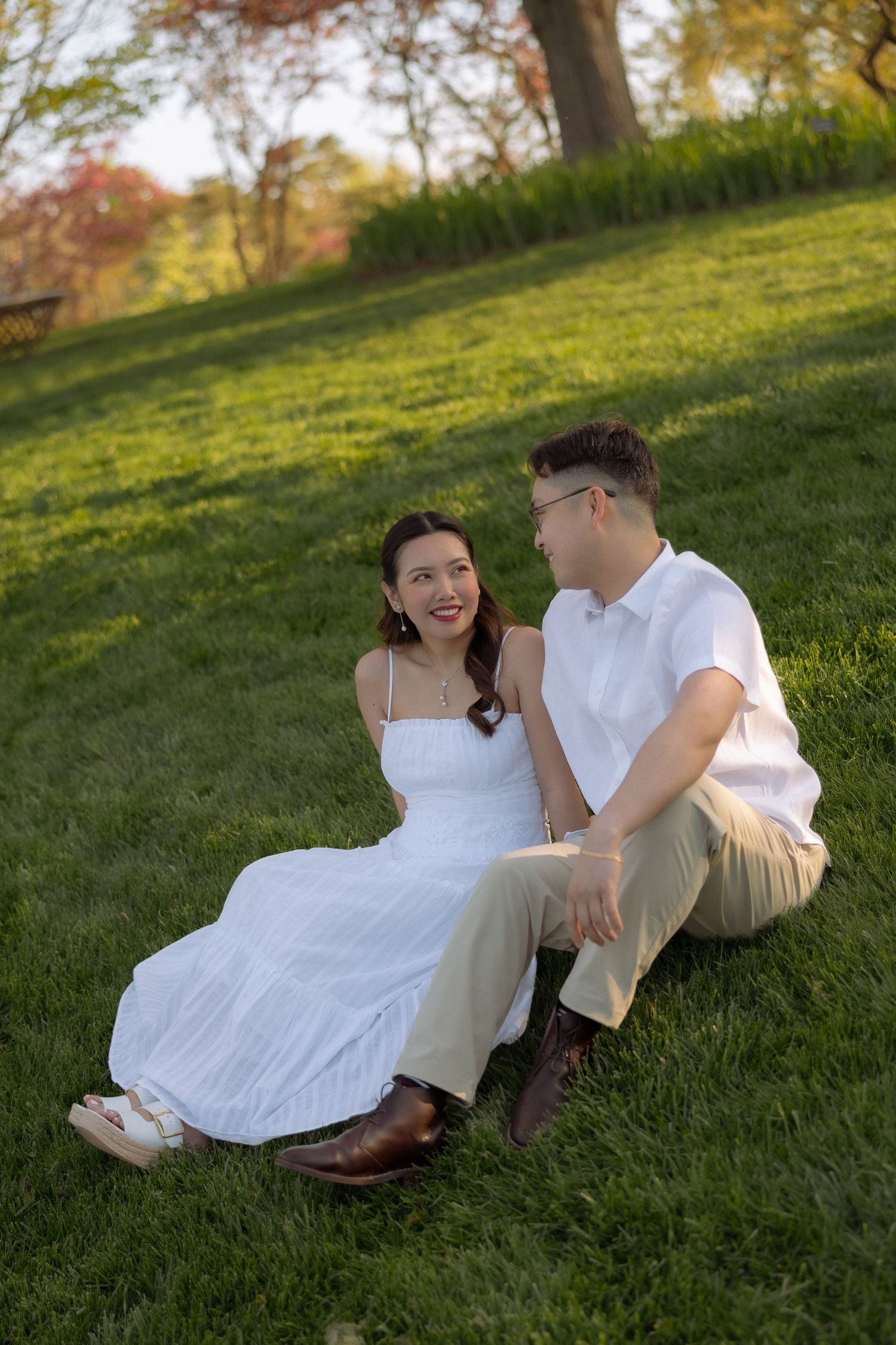 A couple sits on lush green grass in a park, the woman in a white dress and man in white shirt smiling at each other.