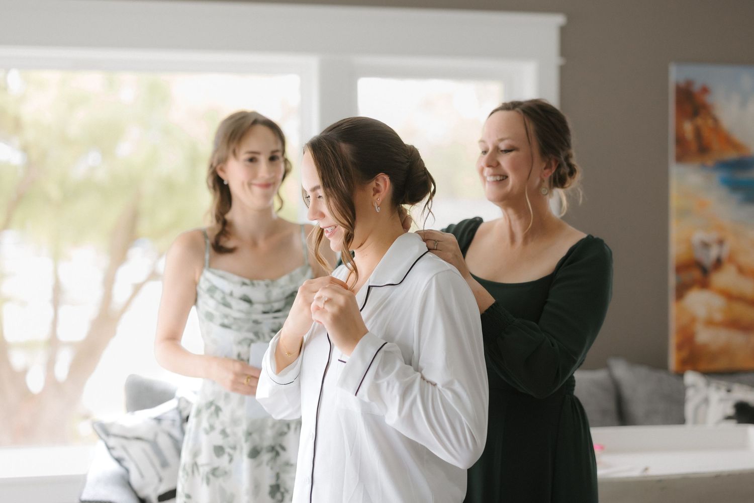 Bride in white satin pajamas laughs with two women helping her get ready on wedding morning indoors.