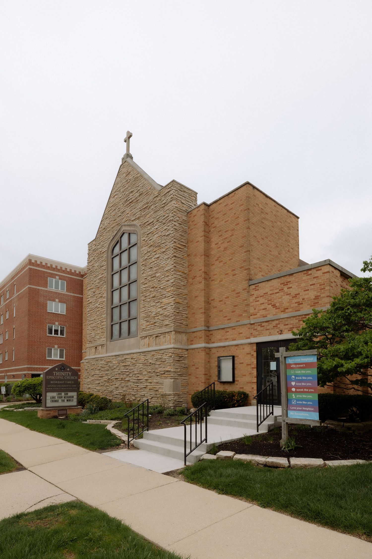 Stone and brick church building with a cross on top, large arched window, and colorful sign out front on a cloudy day.