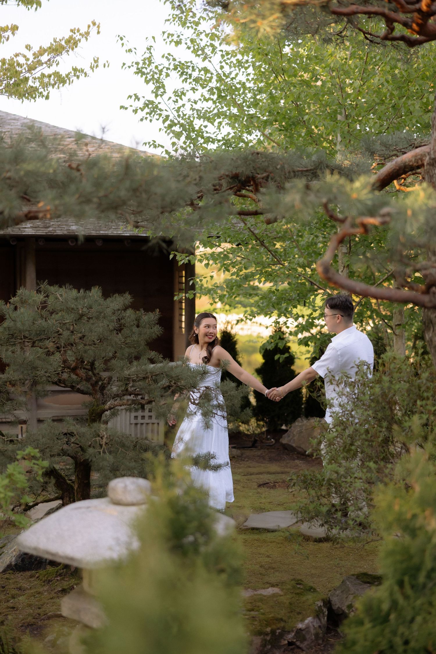 Couple in white holding hands in a serene Japanese garden with lush green trees and a traditional building backdrop.