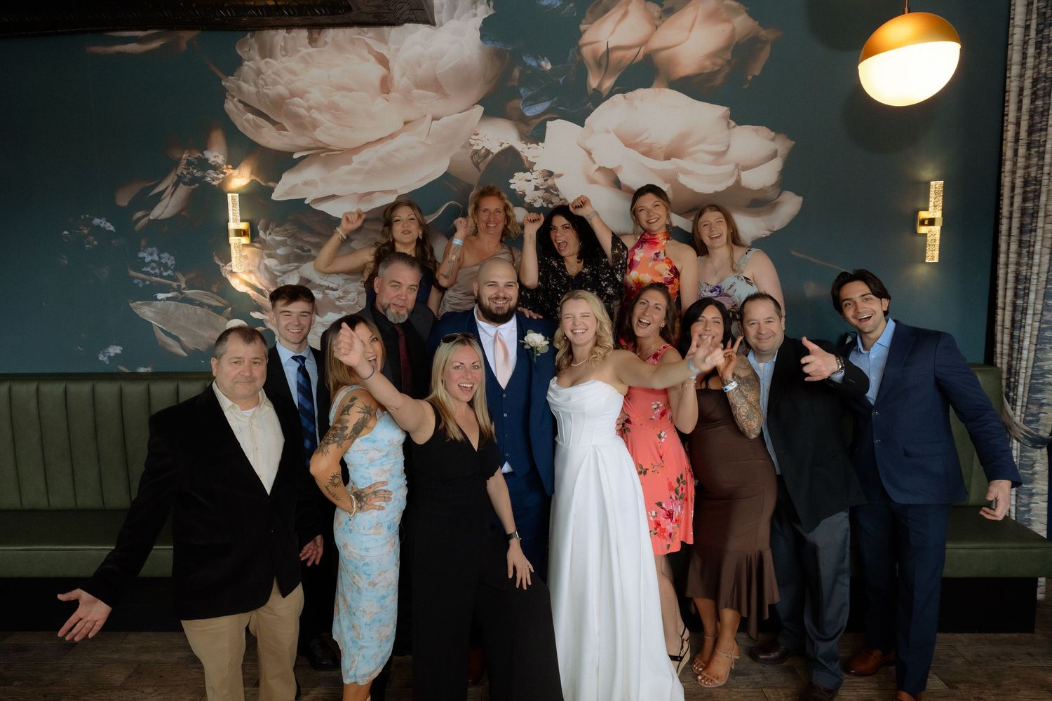 Wedding couple in white poses with smiling guests in front of floral wall decor at reception venue.