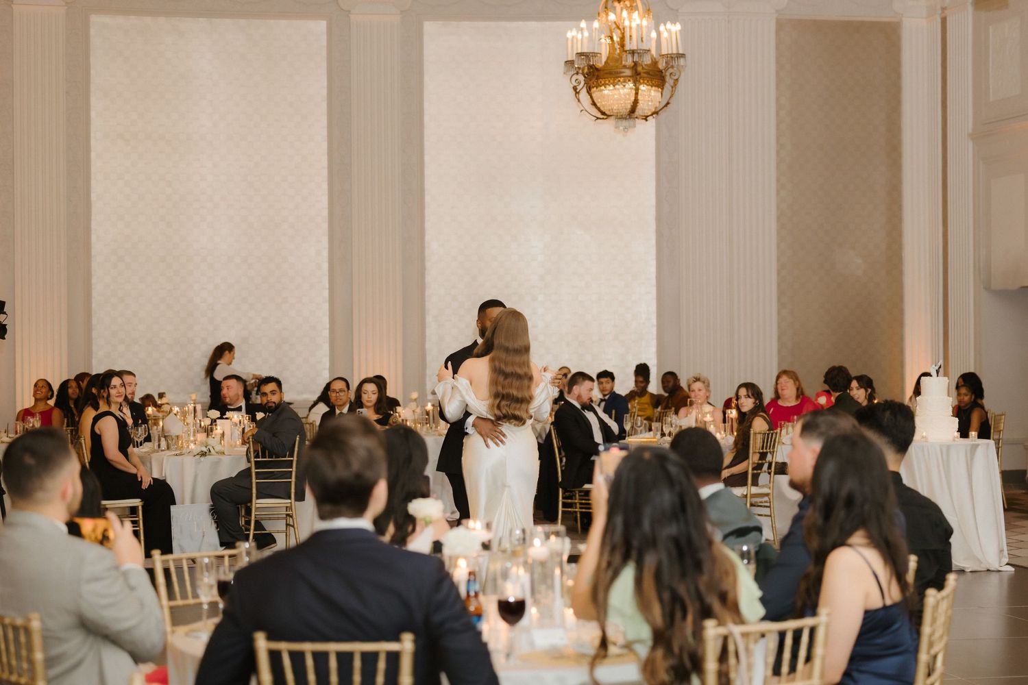 Bride and groom share first dance in elegant ballroom with guests seated at decorated tables and chandelier above.