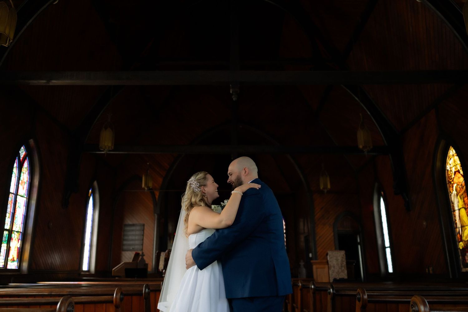 Bride and groom share a romantic embrace inside a historic church with stained glass windows on their wedding day.