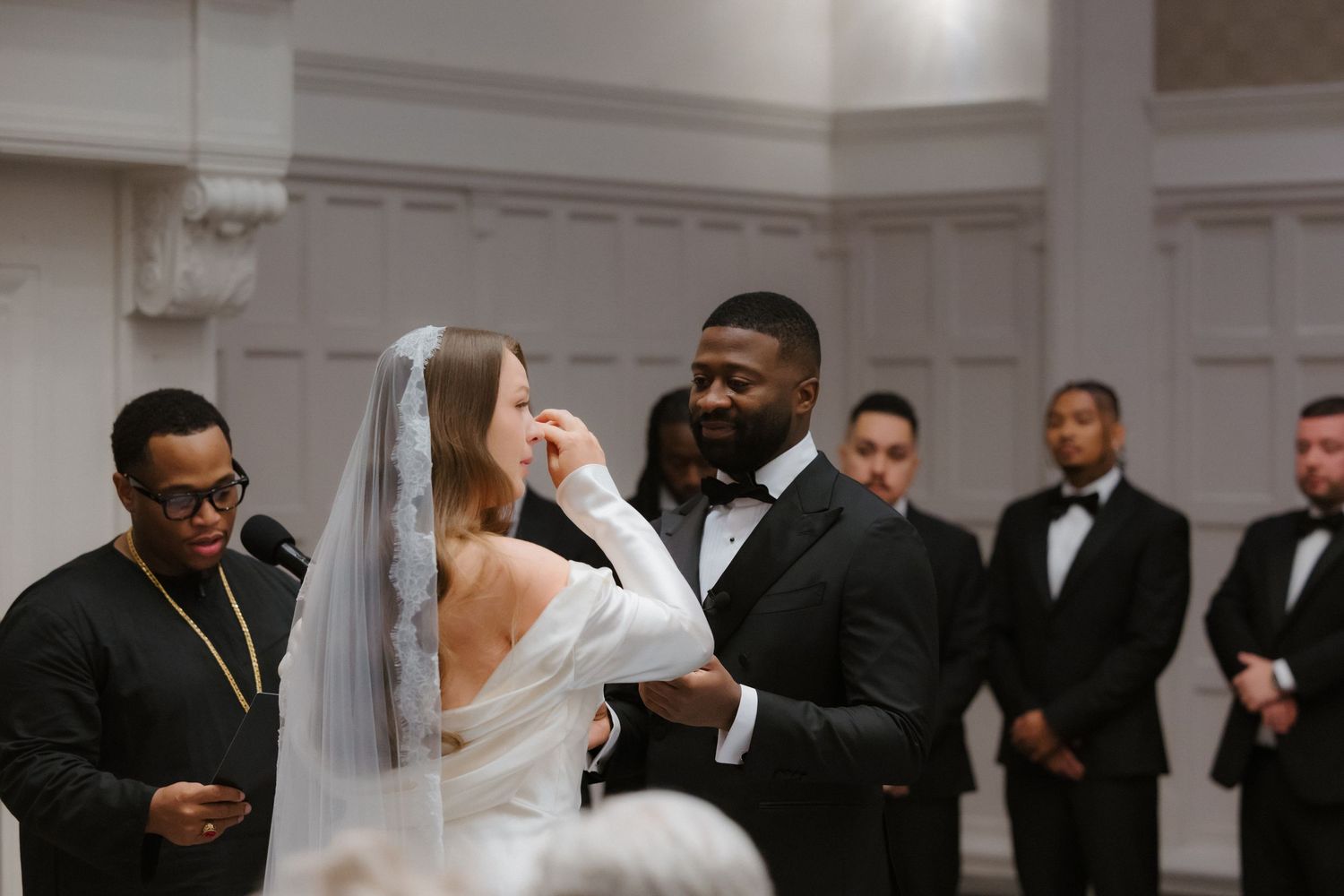 Bride and groom exchange vows at elegant wedding ceremony with groomsmen standing behind them in black tuxedos.