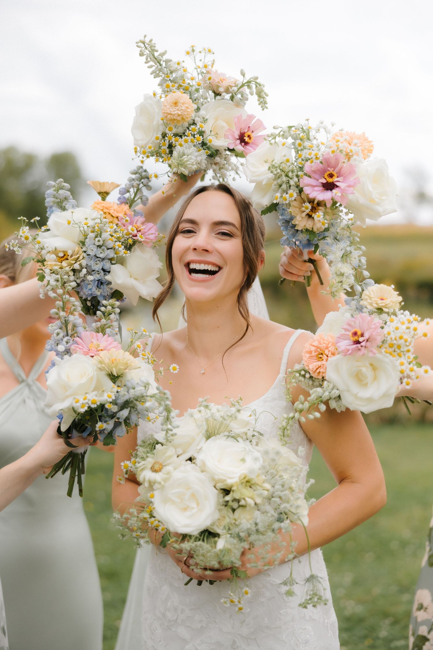Laughing bride in lace gown holding white bouquet, framed by colorful floral arch held by bridesmaids outdoors.