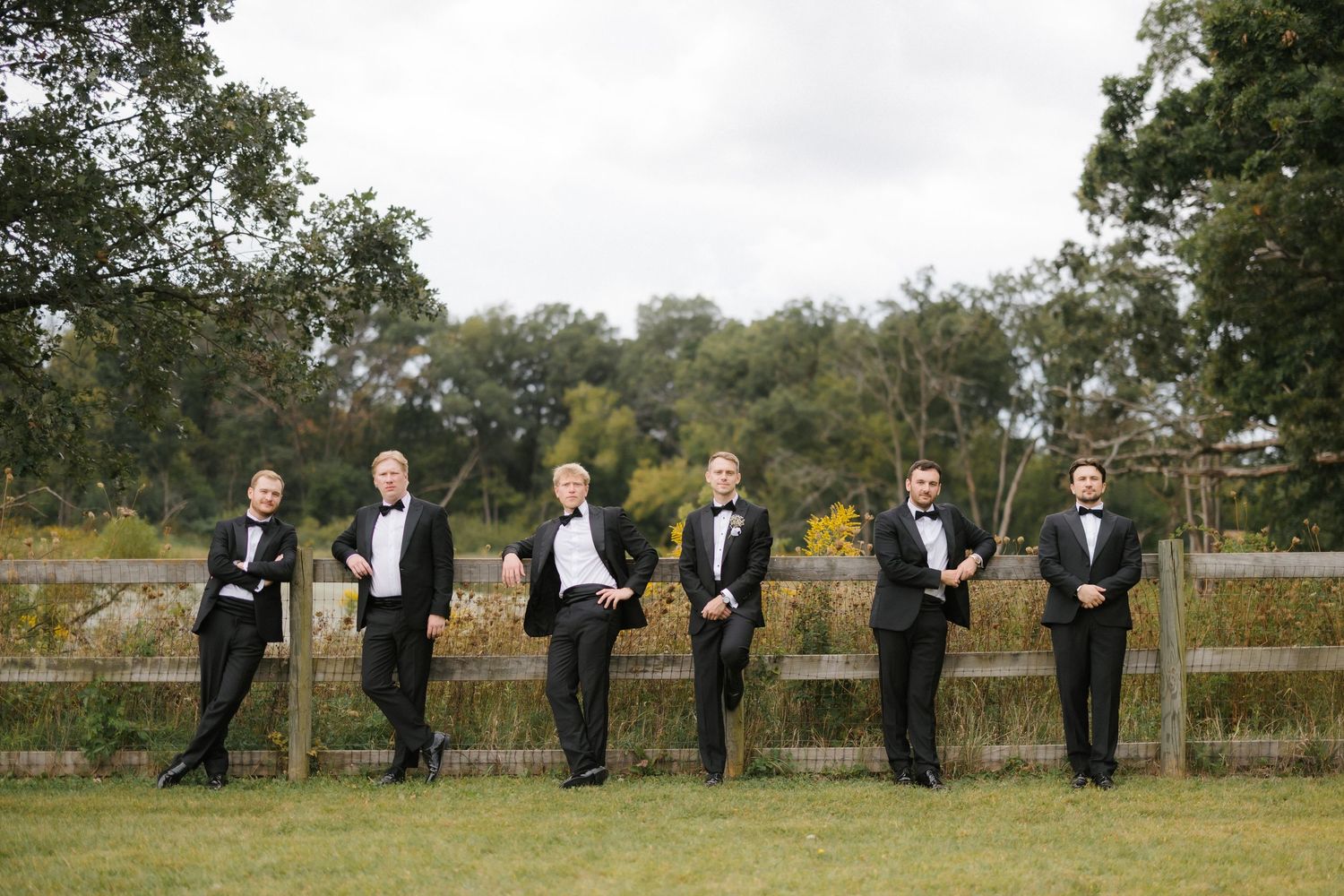 Six groomsmen in classic black tuxedos lean casually against a wooden fence in a lush green outdoor setting.