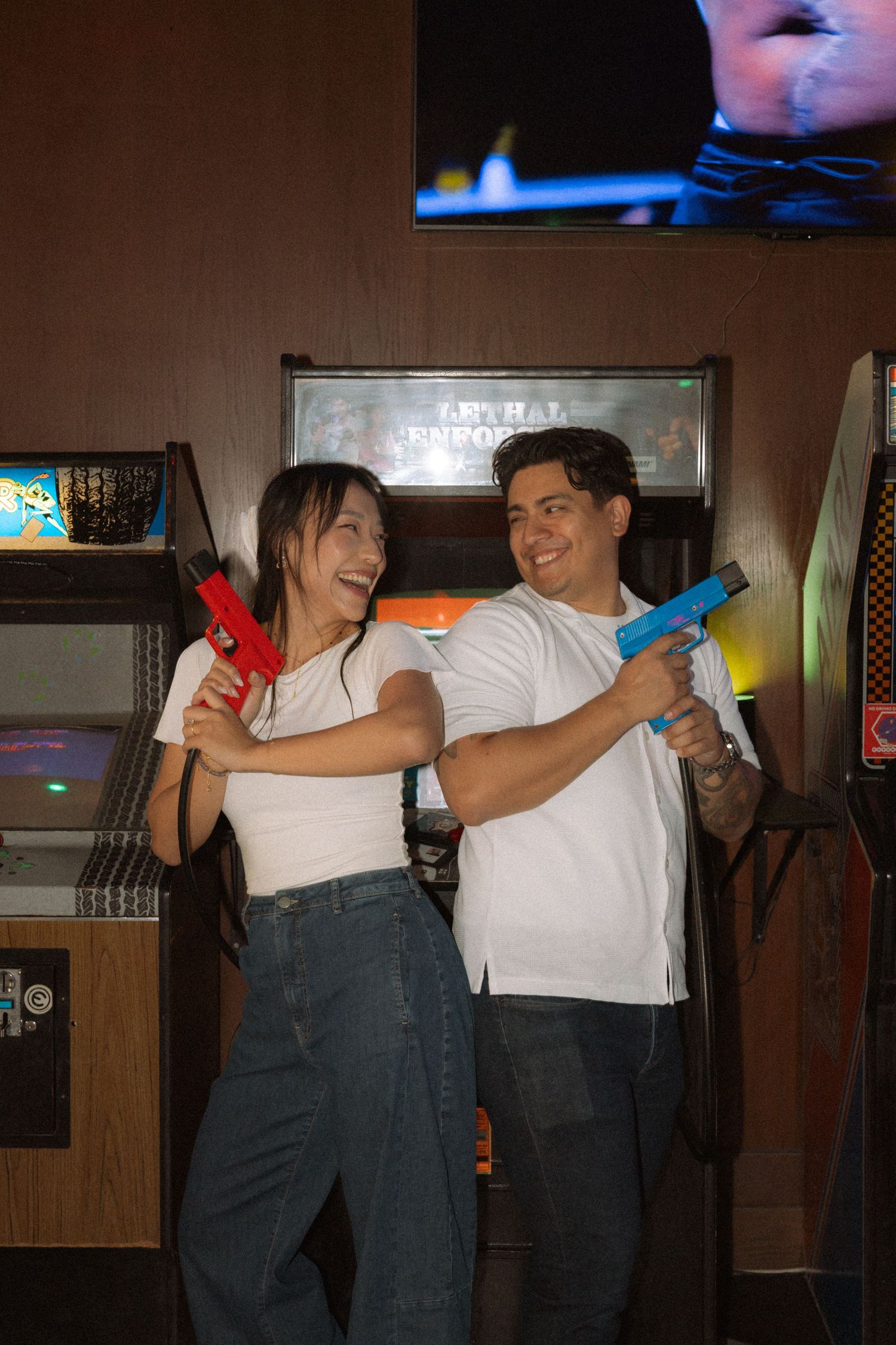 Smiling couple posing with colorful toy guns in front of Lethal Enforcers arcade machine.