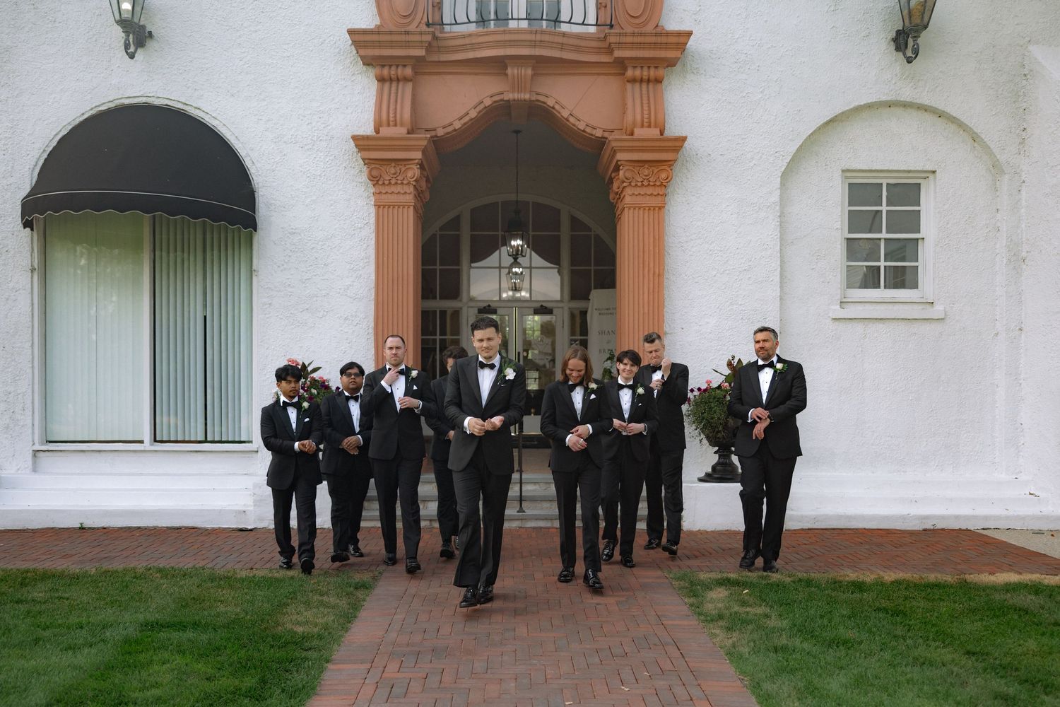 Groom leads groomsmen in black tuxedos outside elegant orange and white venue entrance.