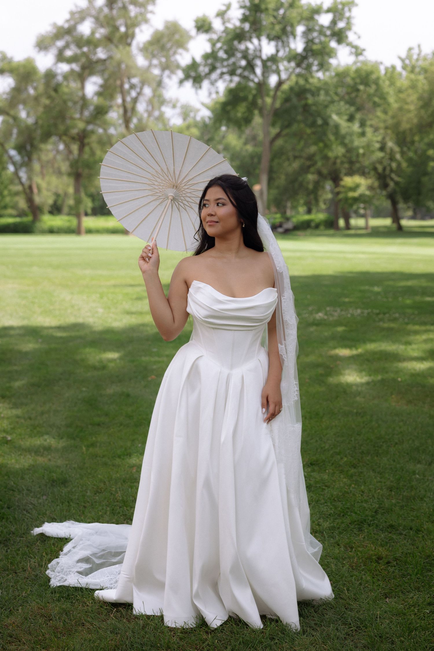 Bride in strapless white ball gown holds white parasol umbrella while posing in green park.