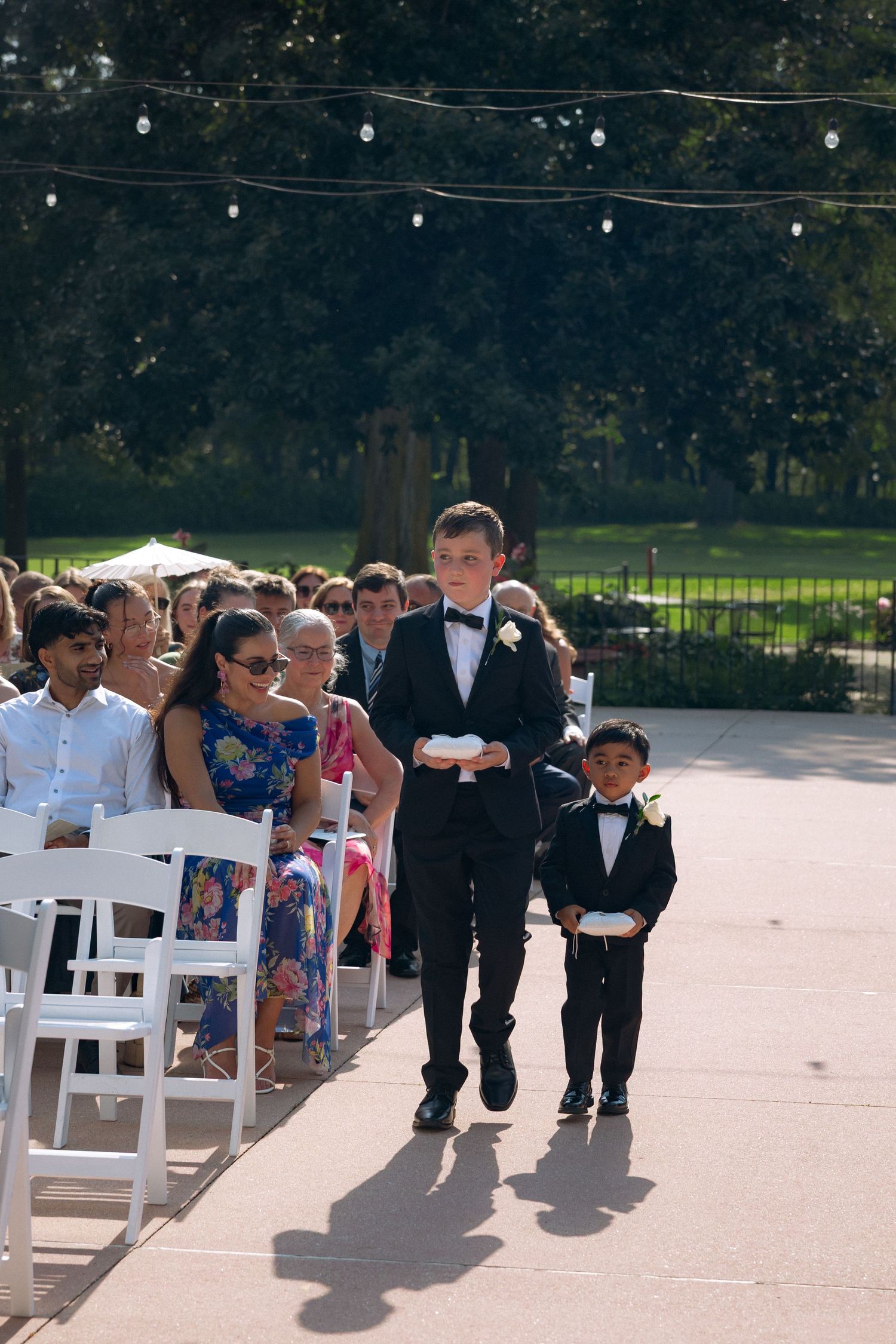 Two young boys in tuxedos walk down outdoor wedding ceremony aisle as ring bearers.
