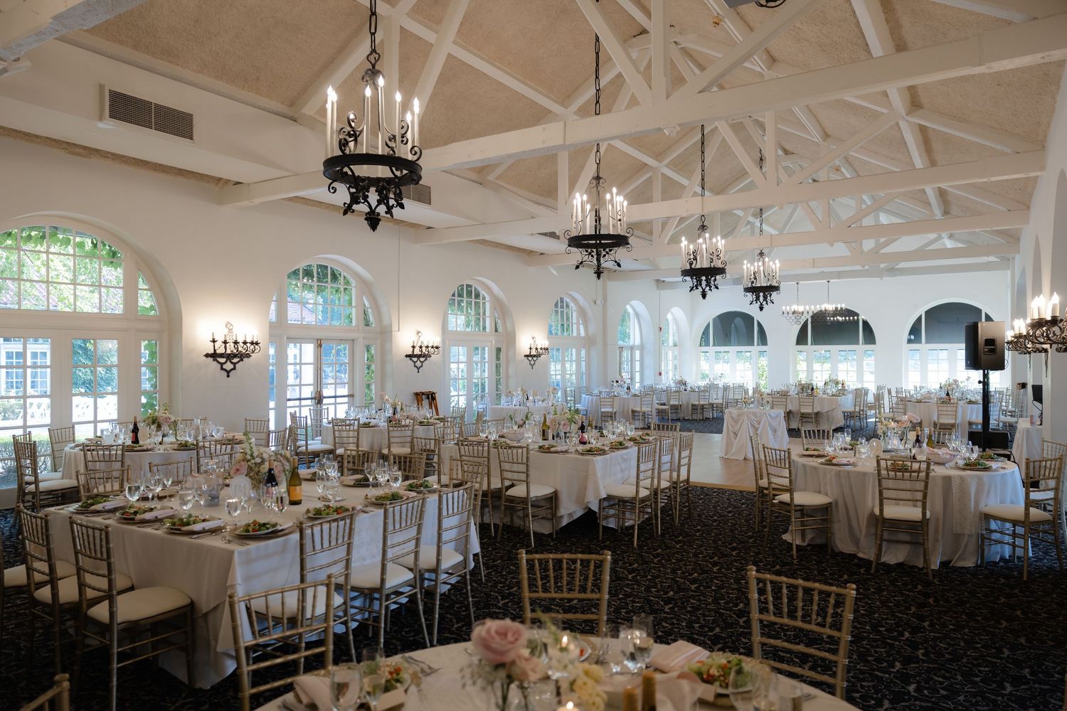 Elegant wedding reception hall with white vaulted ceilings, chandelier lighting, and gold chiavari chairs.