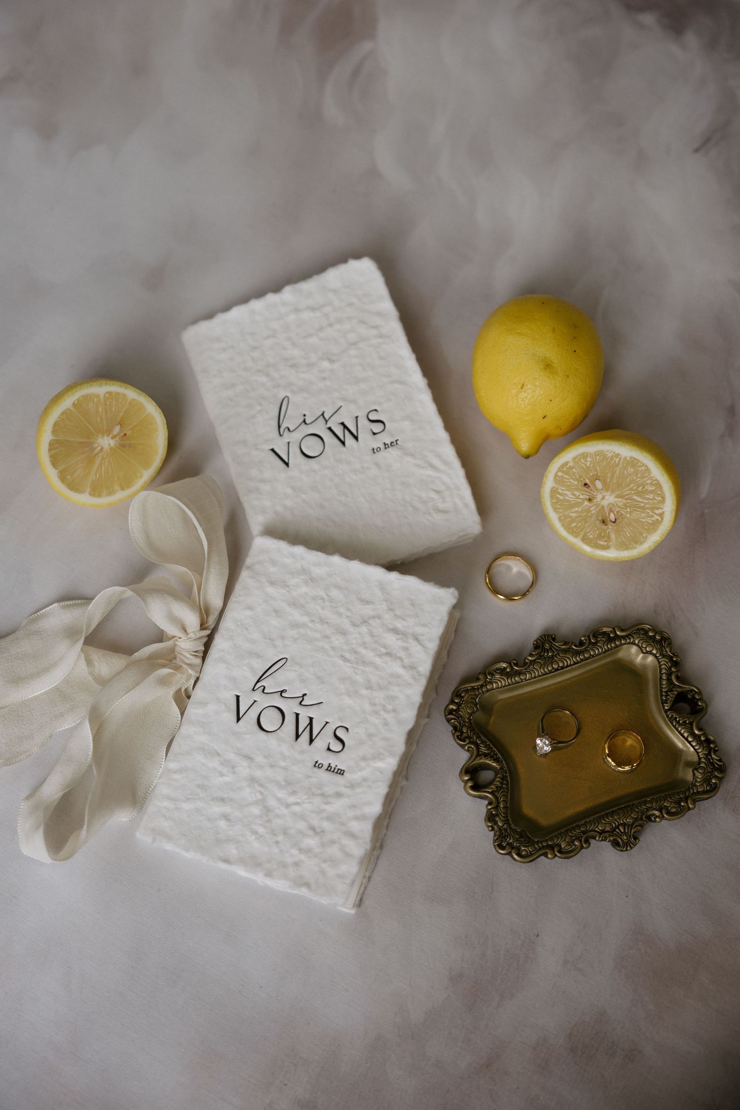 Wedding vow books labeled his and hers displayed with gold rings on ornate tray, fresh lemons, and ivory ribbon on white fur.