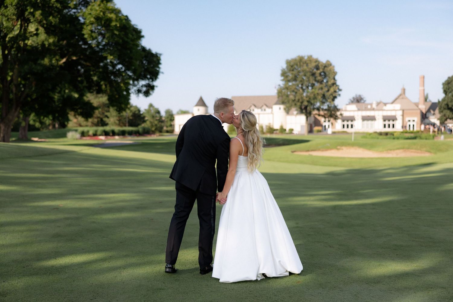 Bride and groom kissing on golf course with elegant country club building in background.