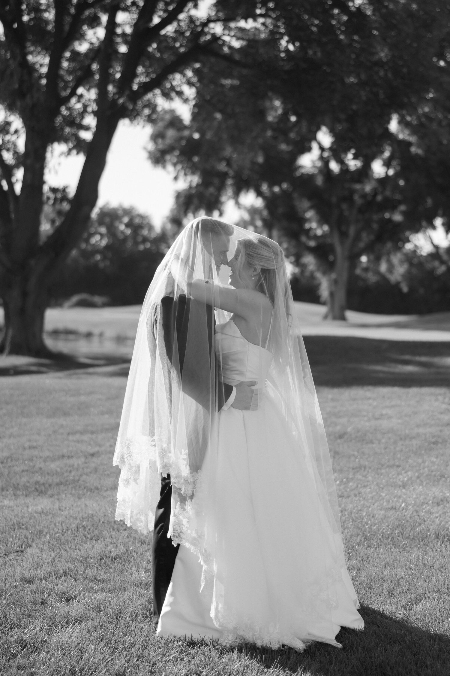 Bride and groom share a romantic kiss under her veil in a black and white outdoor portrait.