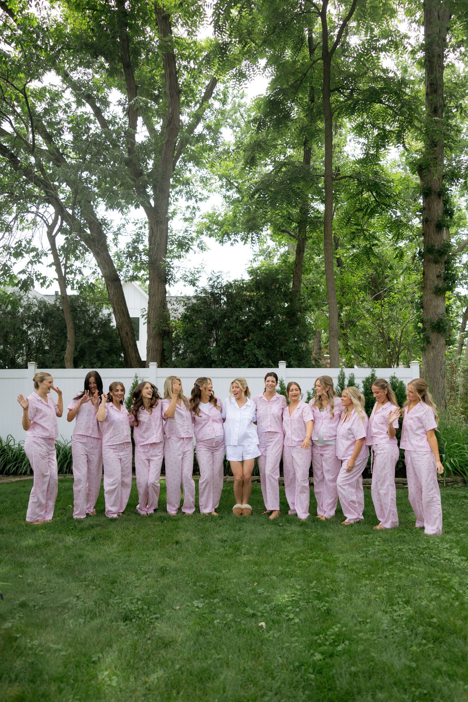 Bride in white robe poses with bridesmaids in matching pink pajamas on green lawn with trees.