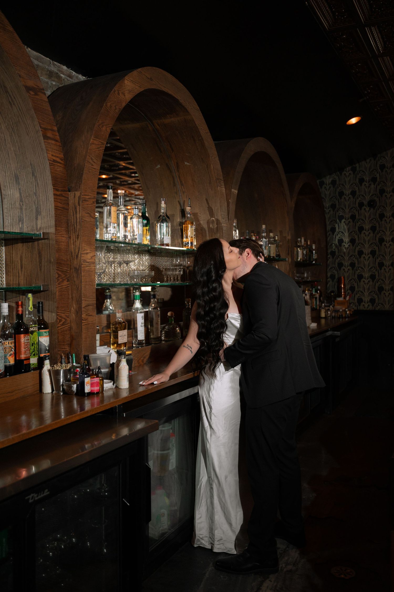 Bride and groom share a romantic kiss at a dimly lit bar with arched wooden shelving and bottles.