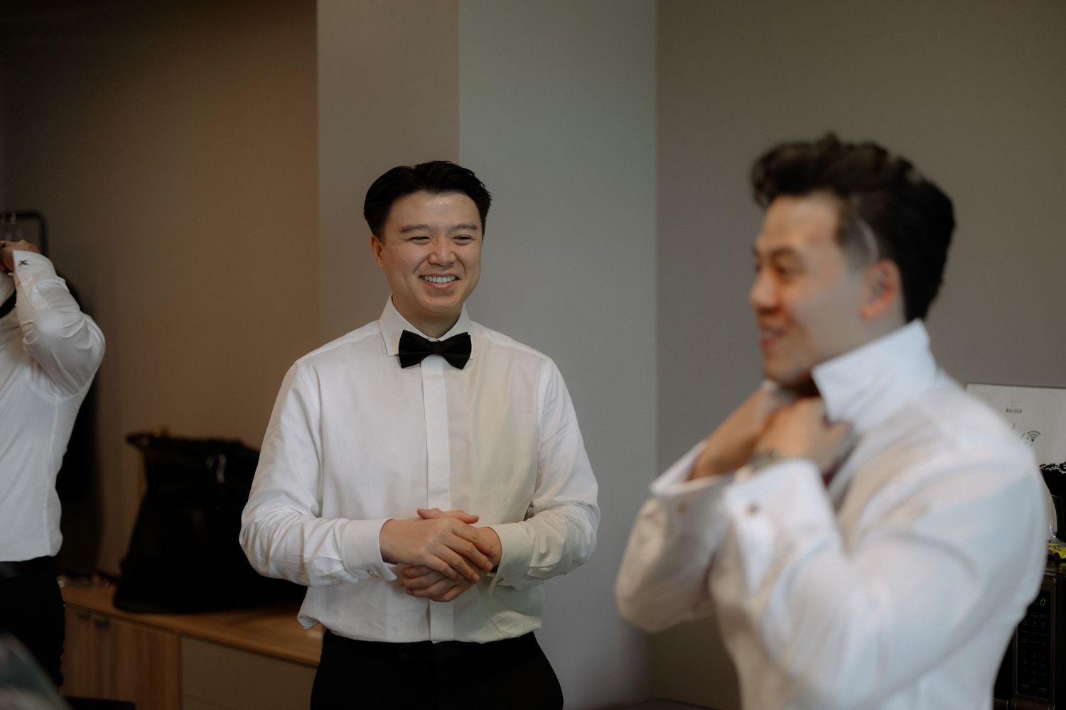 Two groomsmen in white dress shirts and bow ties smiling and adjusting ties while getting ready for a wedding.