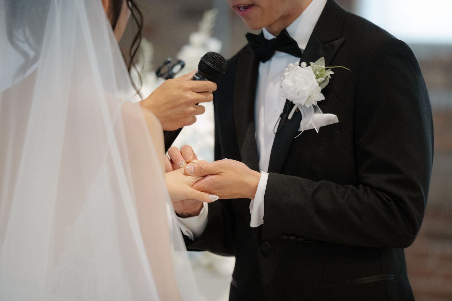 Groom placing wedding ring on bride's finger during ceremony, wearing black tuxedo with white boutonniere.