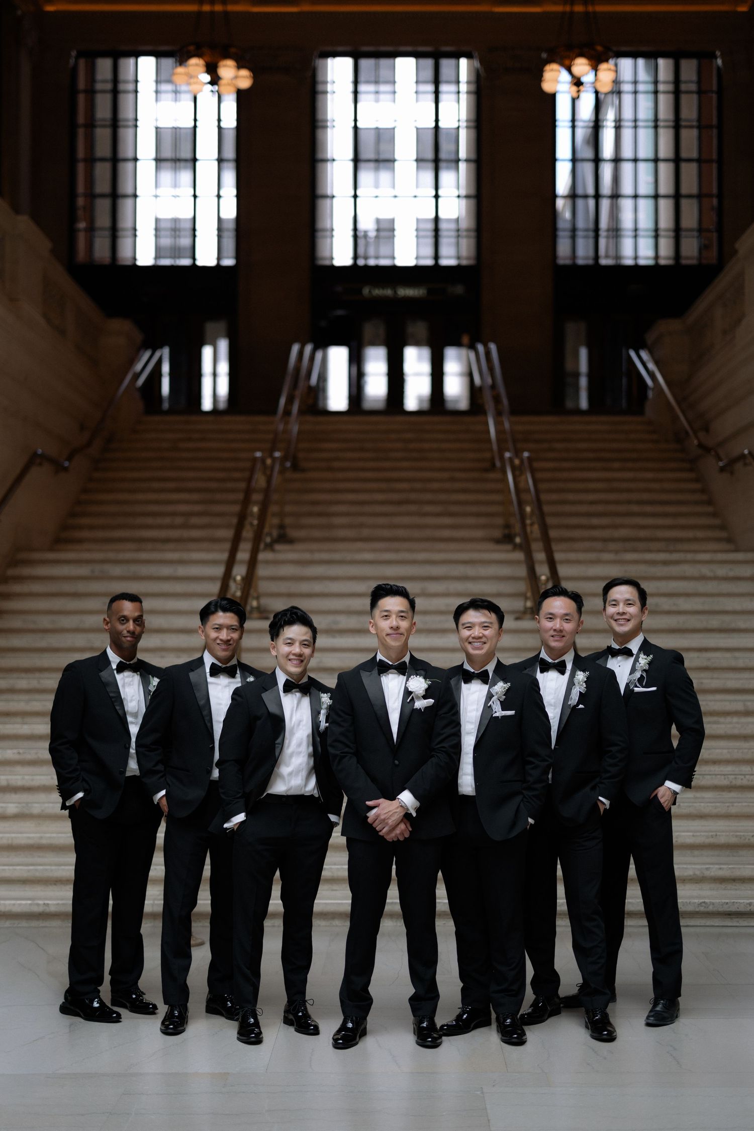 Seven groomsmen in black tuxedos pose together inside elegant hall with grand staircase backdrop.