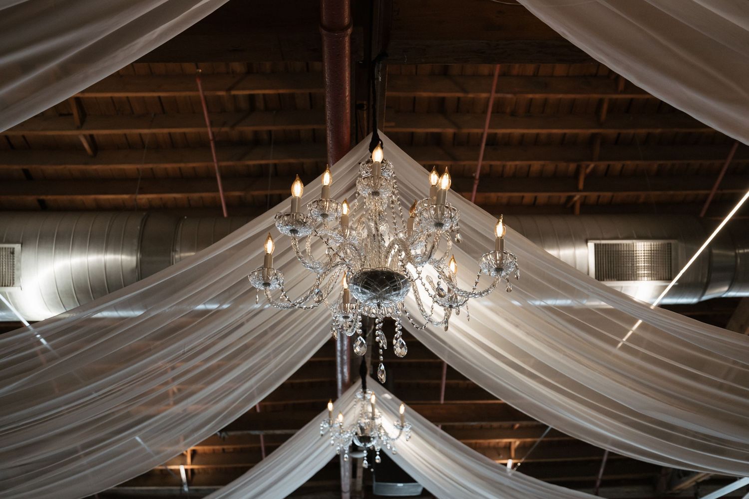 Crystal chandelier hanging from draped white fabric ceiling in rustic barn wedding venue with exposed wooden beams.