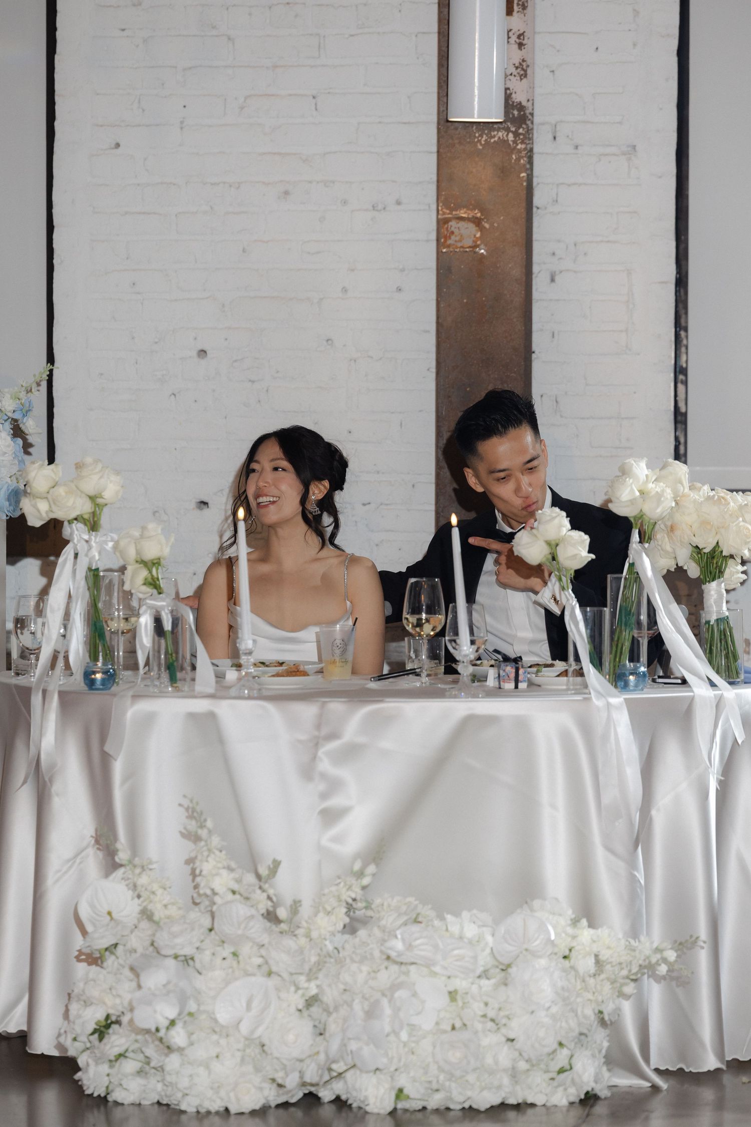 Bride and groom smiling at sweetheart table adorned with white floral arrangements.