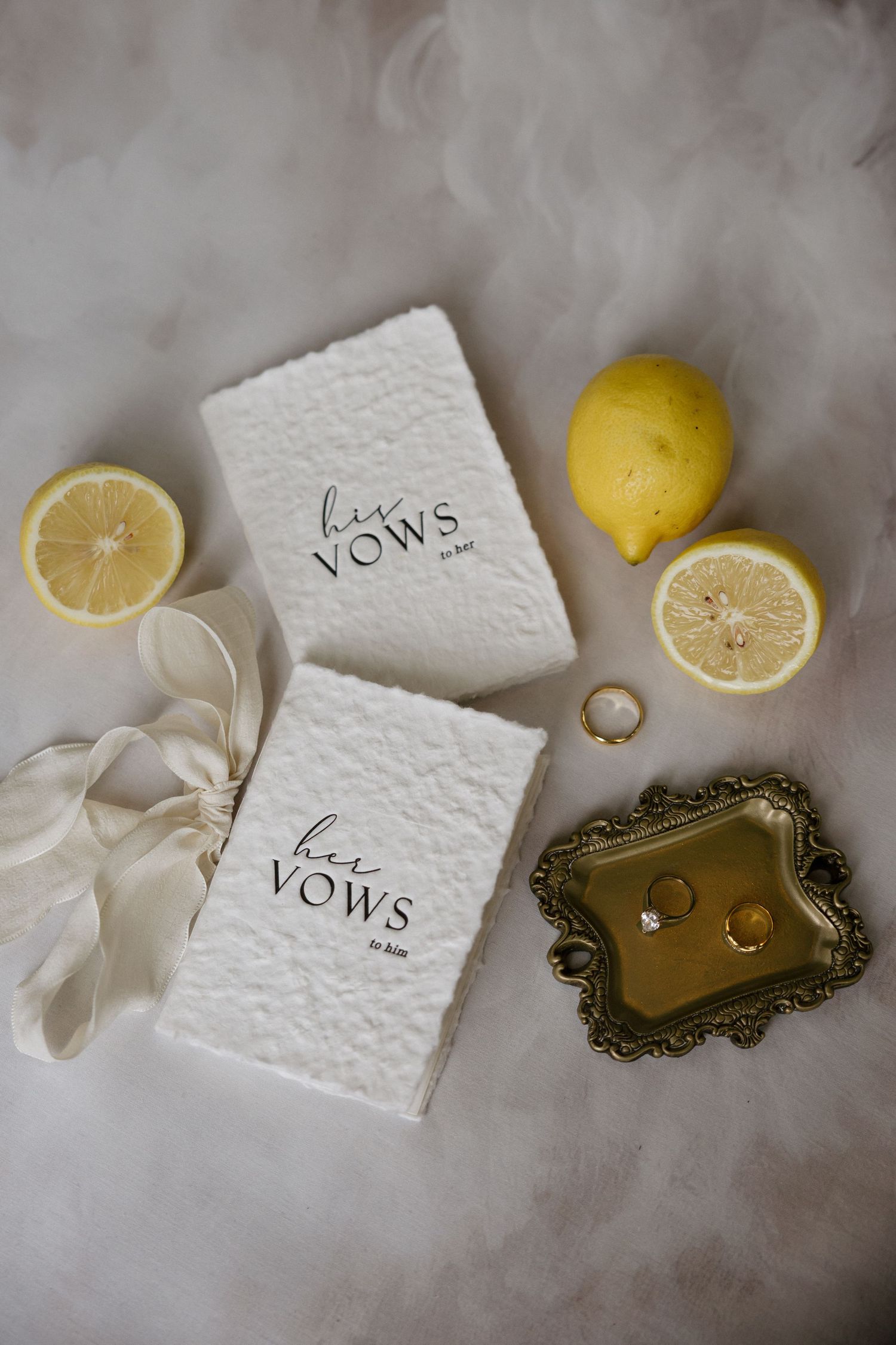 Wedding vow books, gold rings on ornate tray, lemons, and ribbon on soft white background.