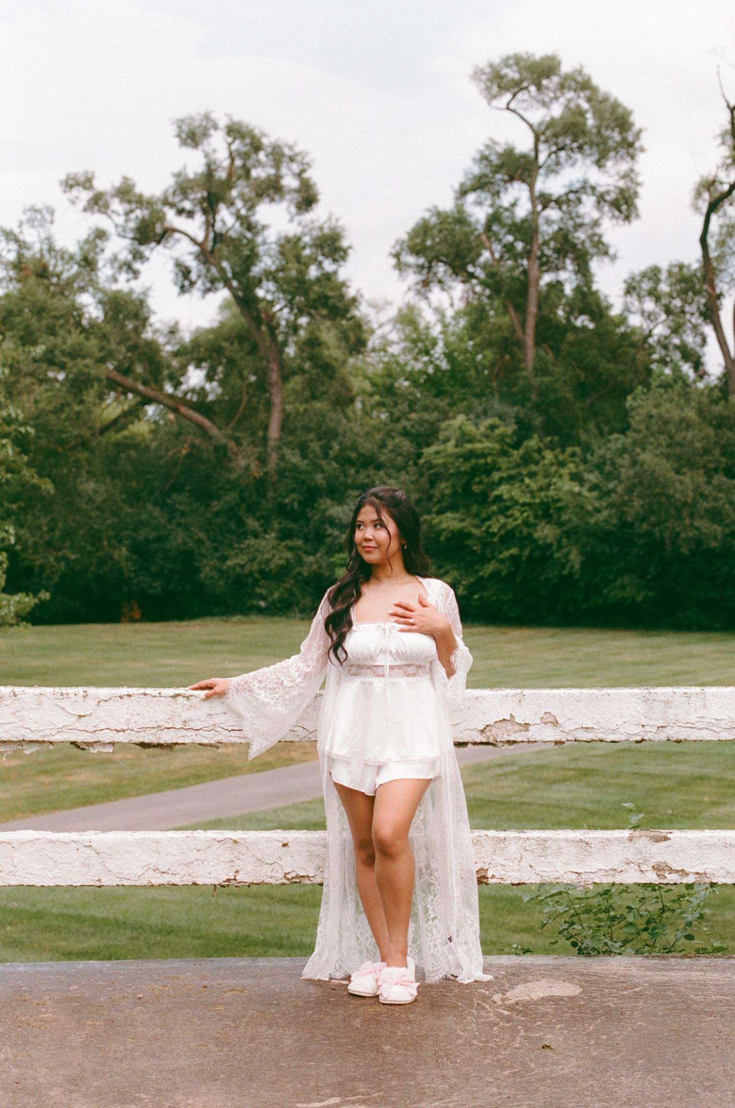 Woman in white lace maxi robe and romper poses outdoors near white fence with lush green trees behind her.