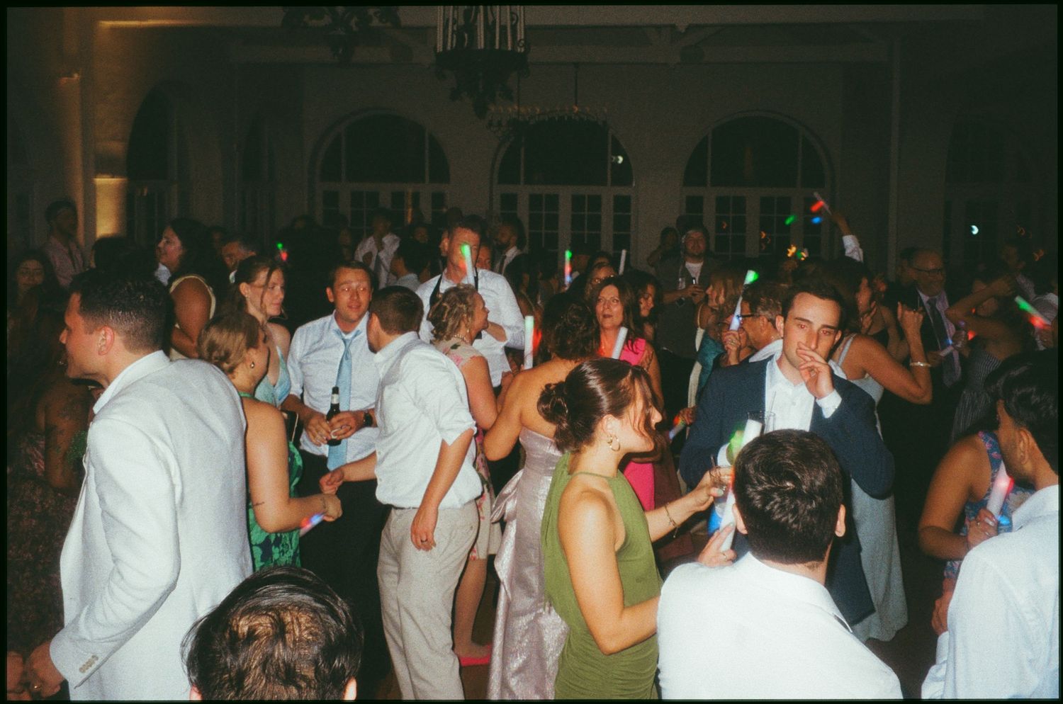 Crowd of elegantly dressed guests dancing at a formal wedding reception in a grand ballroom venue.