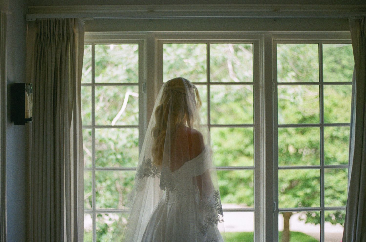 Bride in wedding gown and veil standing before large window overlooking lush green garden on wedding day.